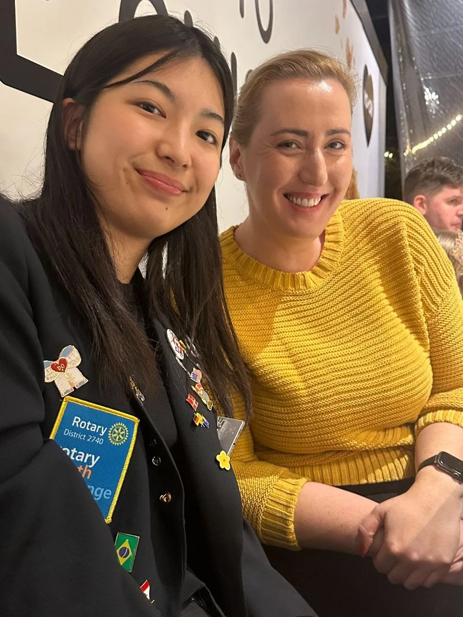 Two women sitting next to each other, smiling at the camera. The woman on the left has black hair and is wearing a blazer with various pins, including a Rotary badge. The woman on the right has blonde hair and is wearing a yellow sweater. In the back