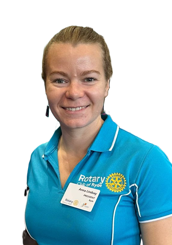 A woman smiling, wearing a blue Rotary Club of Rye polo shirt with a name tag that says Anna Lindsay, President of Rye, standing indoors with other people in the background.