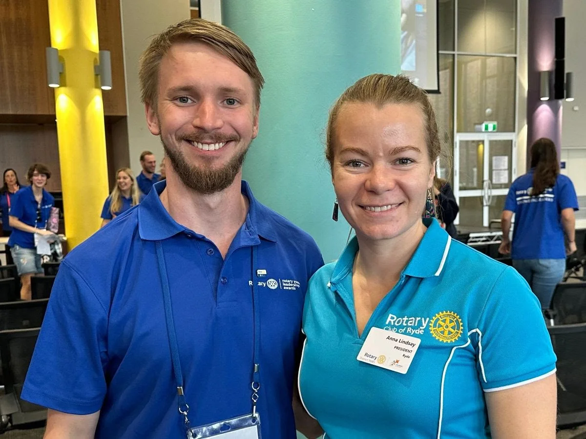 Two smiling individuals, a man and a woman, standing close together at a formal event, wearing blue Rotary Club shirts. The woman has a name badge reading "Anna Lindsay, President." Several other people in yellow and blue shirts are visible in the ba