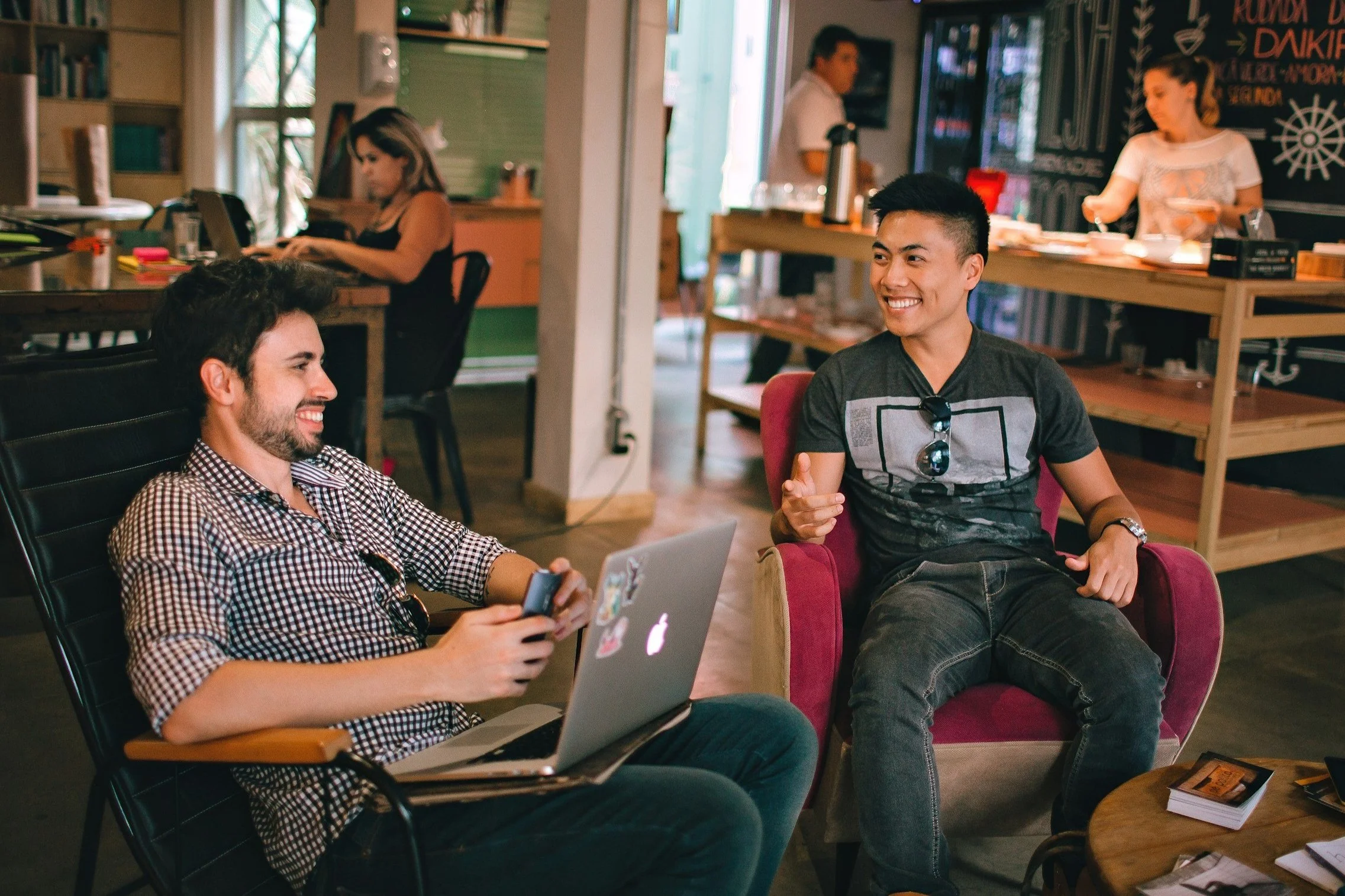 Two young men sitting and talking in a cozy cafe. One is using a laptop and holding a phone, while the other is smiling, wearing sunglasses and gesturing with his hand. In the background, a woman and a man are working or preparing food behind the counter.
