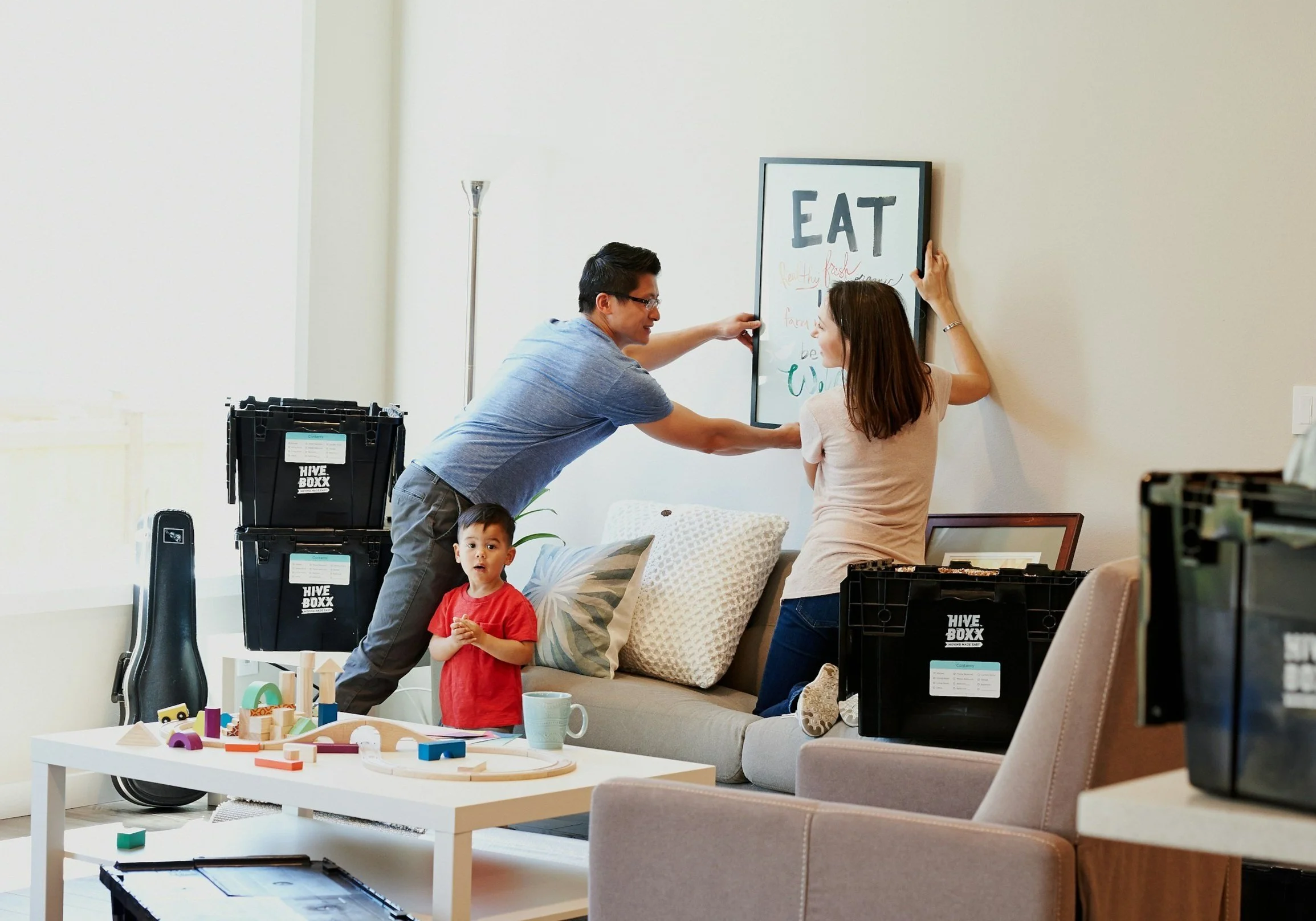 Two adults hanging a framed poster on a white wall in a living room, with a young boy standing nearby, surrounded by moving boxes, toys, and household items.