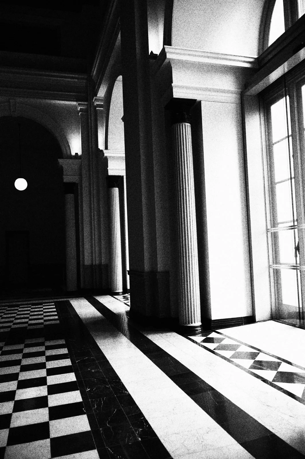 Black and white photograph of an interior space with tall columns, large window, decorative floor tiles, and a door or window with glass panels.