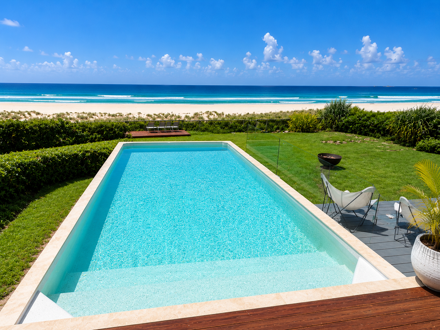 View of a backyard with a clean blue sparkling rectangular swimming pool, green lawn, and an ocean in the background on a sunny day with a blue sky and some clouds.