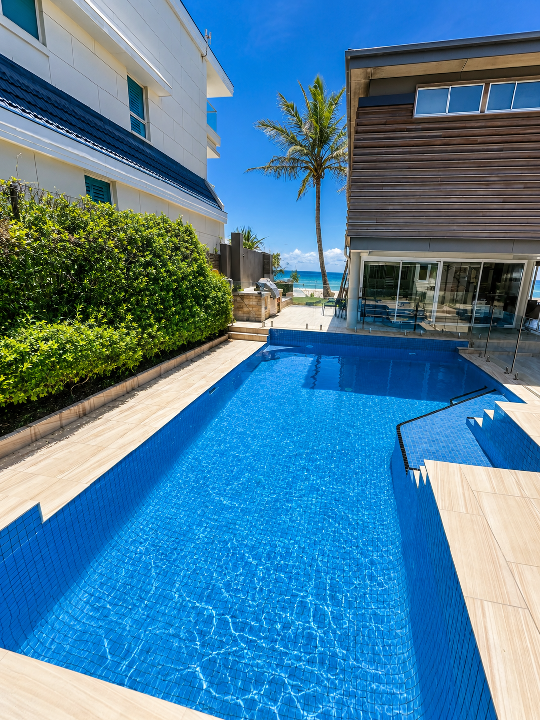 Modern large, clean, sparkling blue swimming pool at a beachfront property with ocean view, modern buildings, and palm trees.