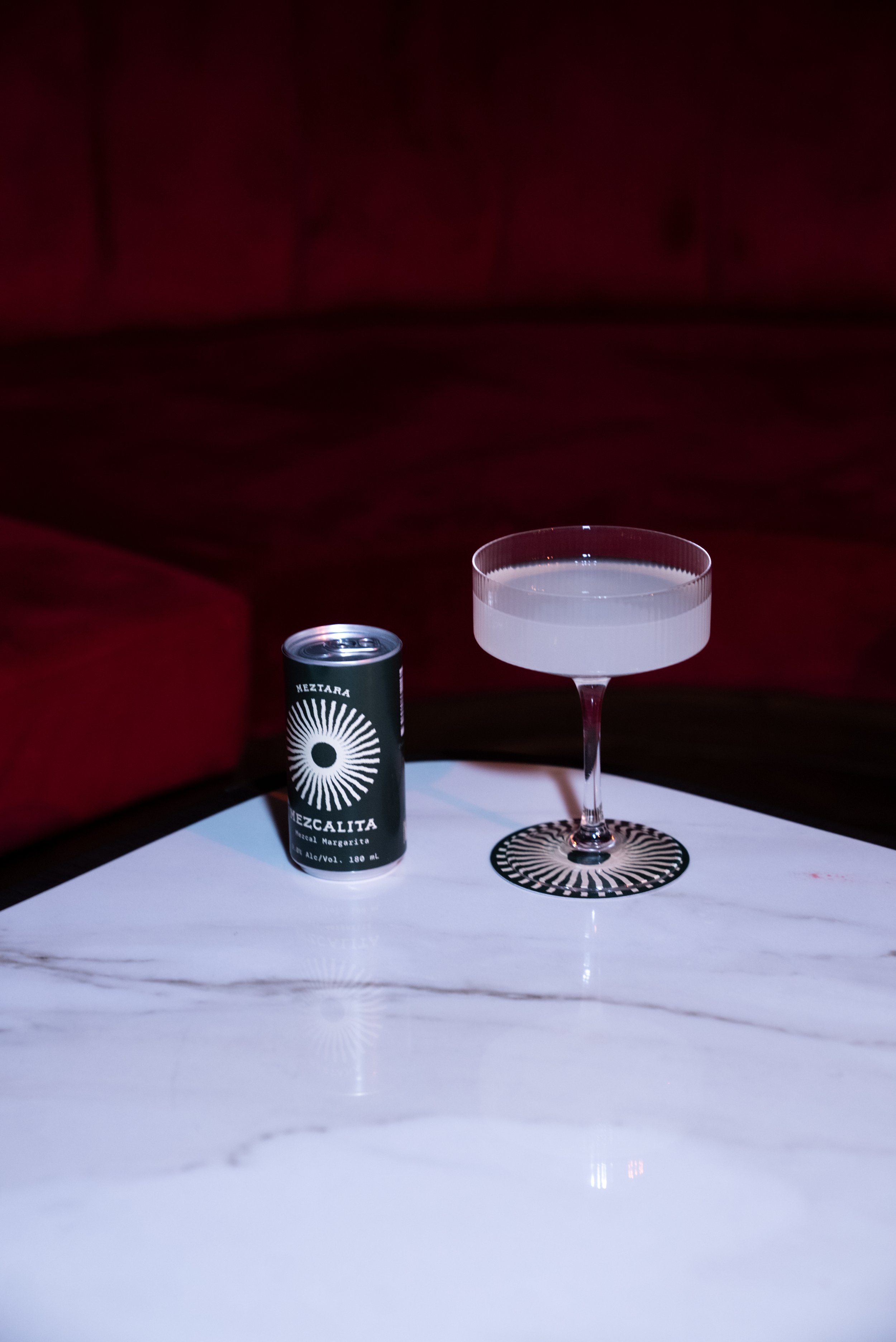 A can of Mezcalita Mexican margarita cocktail next to a coupe glass filled with a light-colored drink on a white marble table with a black and white decorative coaster underneath the glass.