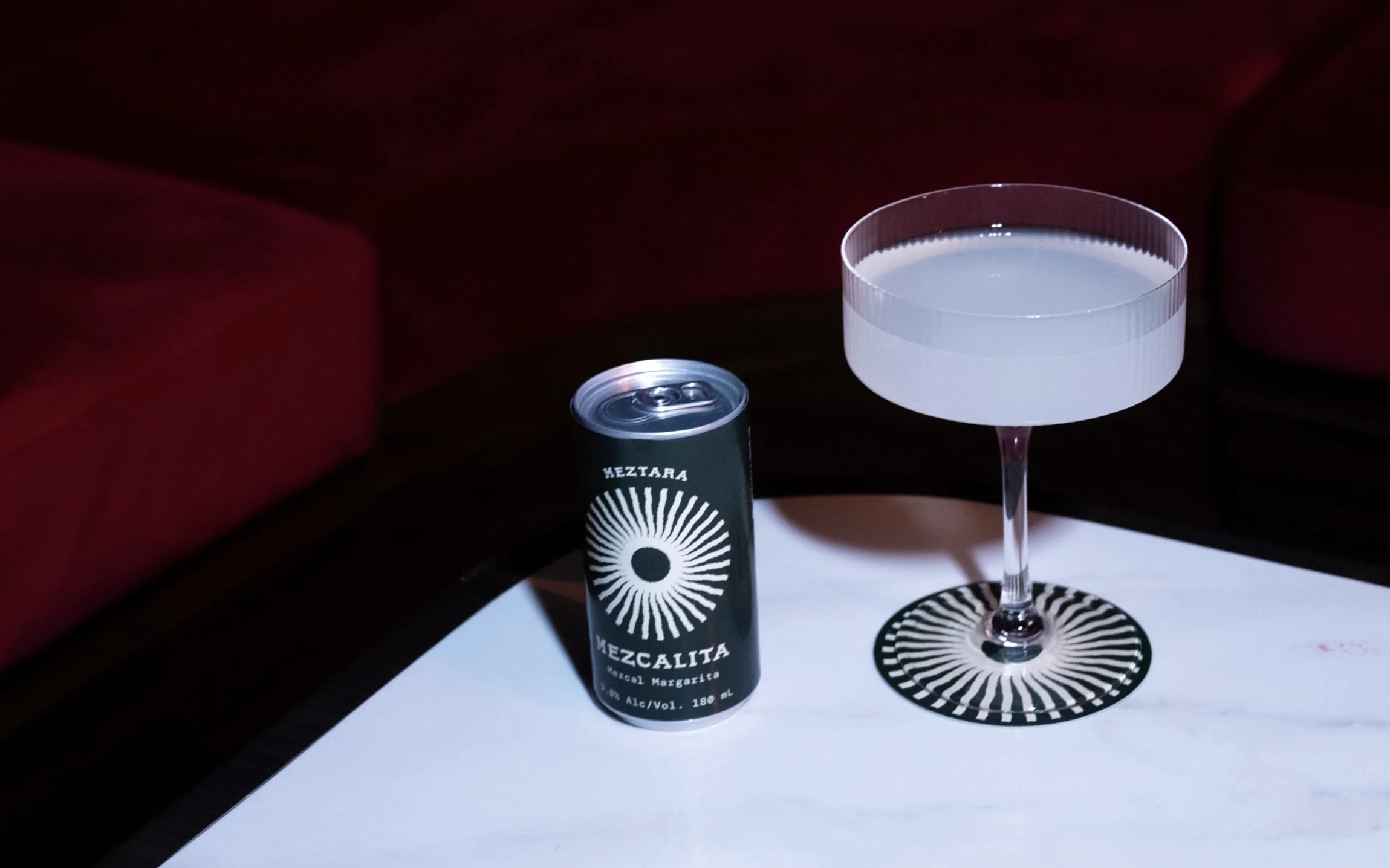A can of Mezcal Margarita cocktail next to a cocktail glass with a clear drink, on a white table with a black and white patterned coaster, with red upholstered furniture in the background.