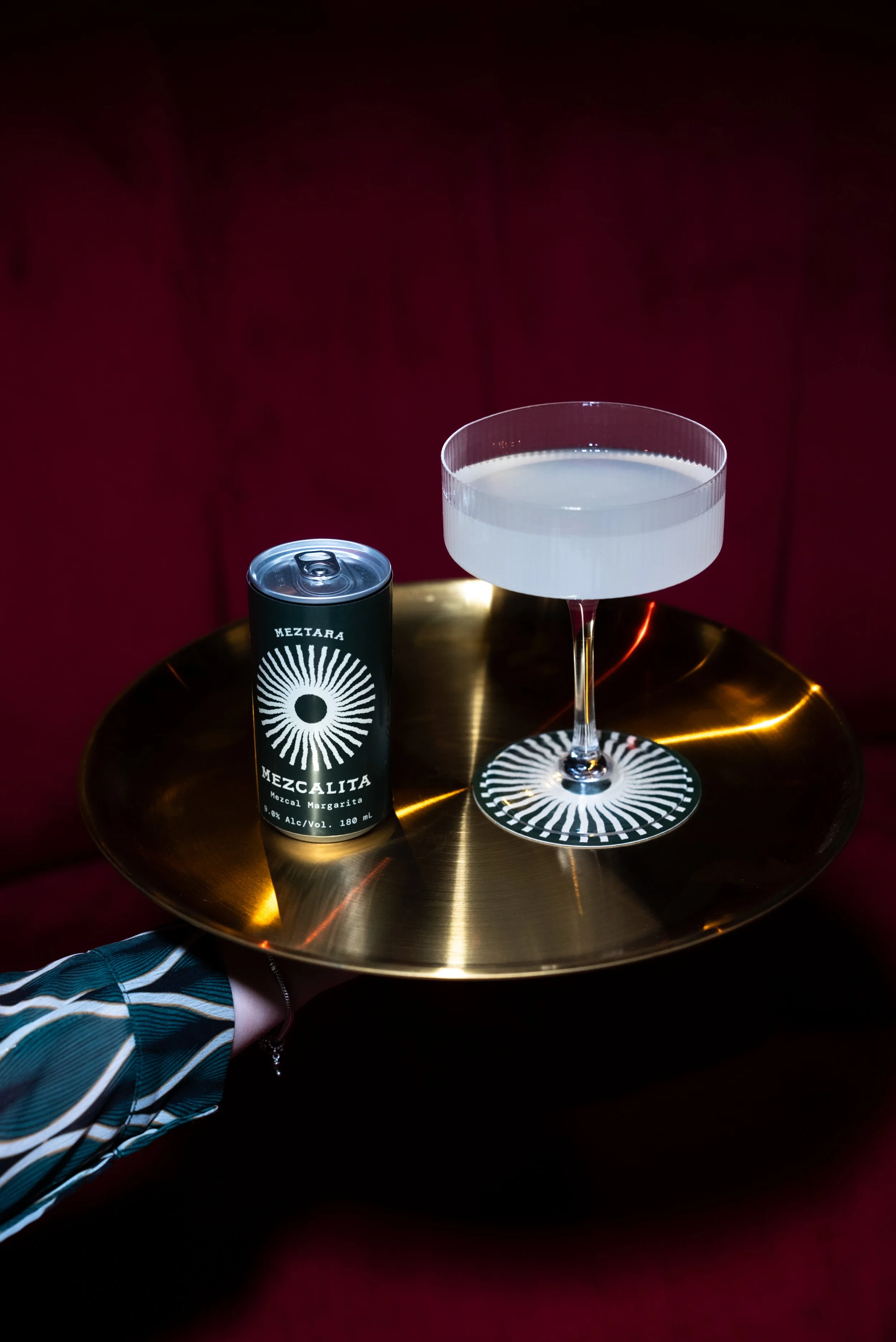 A person holds a gold tray with a black can of Mezcal margarita and a green cocktail glass on it, set against a red background.