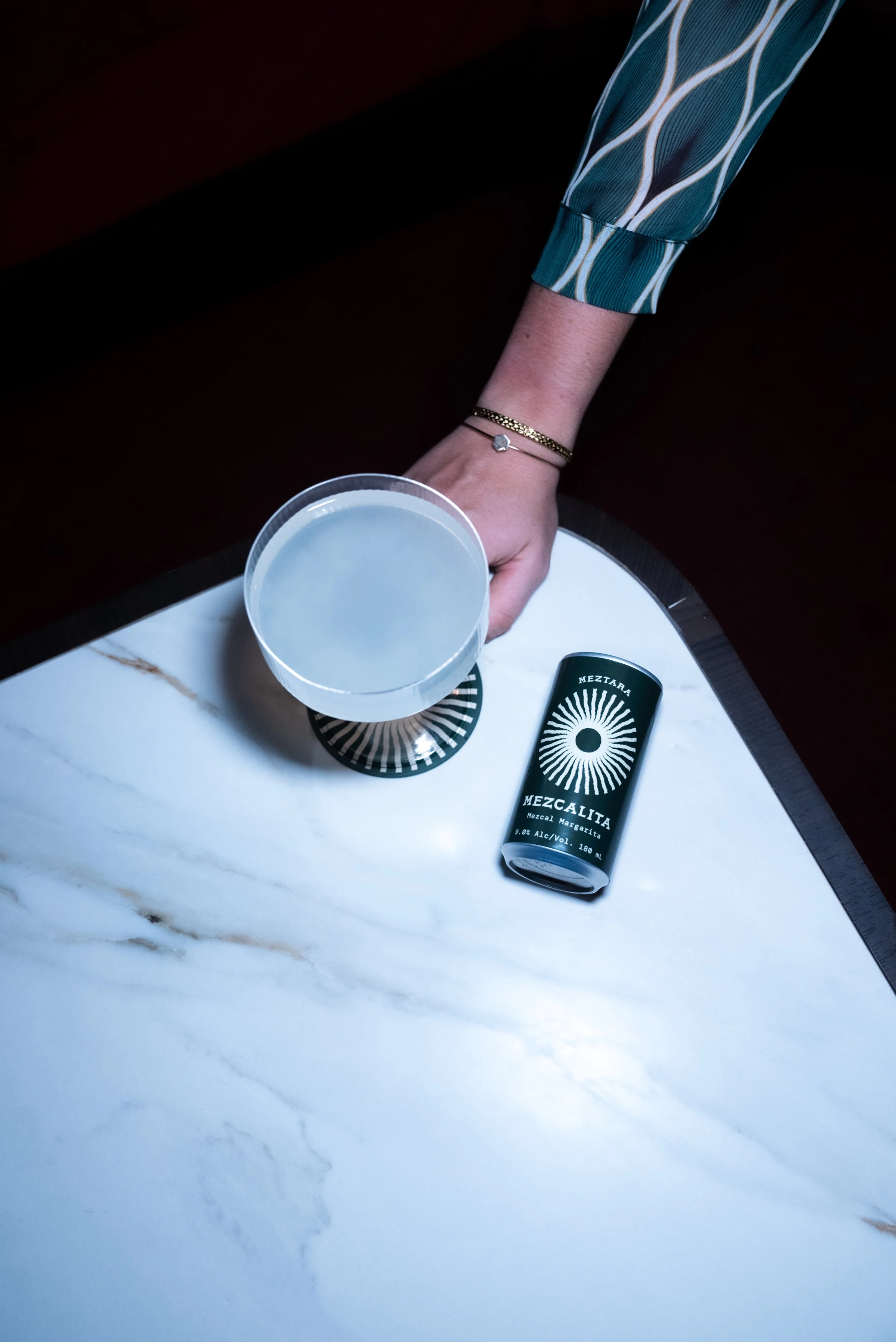 A hand with a gold bracelet holding a cocktail glass with a clear drink, resting on a marble table. Next to the glass is a green can of Mezcal with a black and white sunburst design.