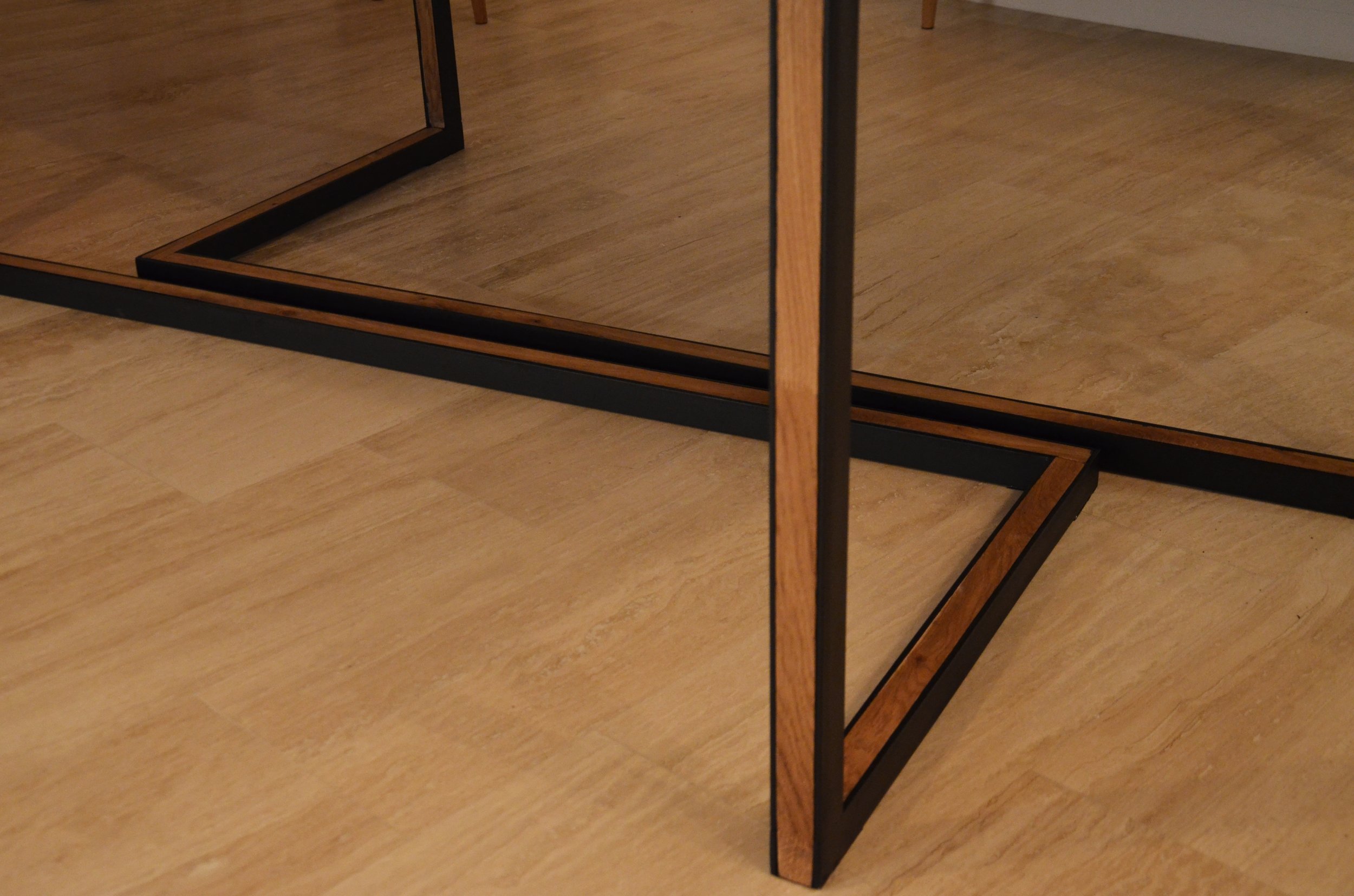 Close-up of a modern wooden and glass table with black metal frame, showing the corner and part of the reflective glass top and wooden leg.