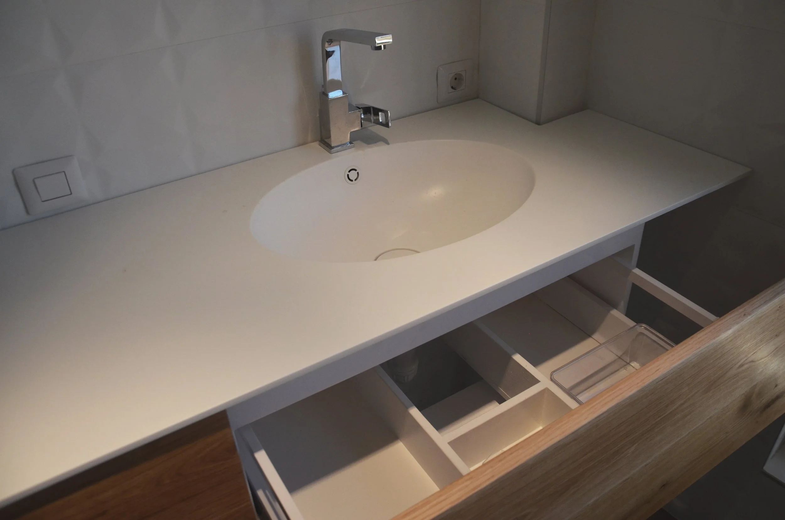 White bathroom sink with a modern chrome faucet, set into a white countertop with a wooden drawer underneath, partly open, revealing interior compartments.