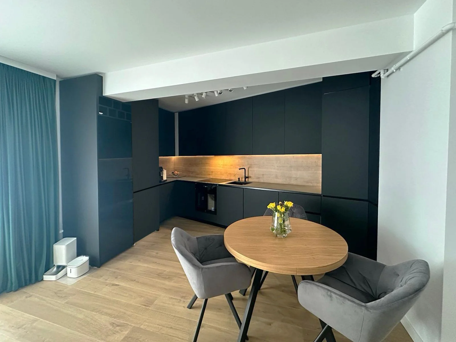 Modern kitchen with dark cabinets, a wooden backsplash, and a small round wooden dining table with two gray upholstered chairs. A small vase of yellow flowers is on the table.