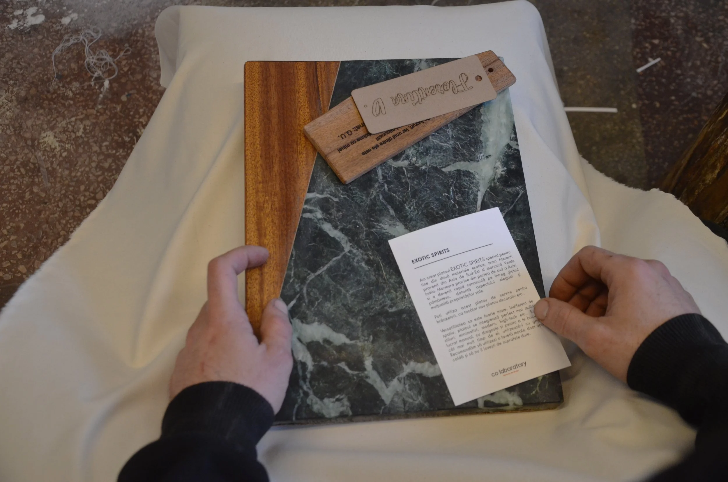 Two hands are holding a black marble tablet with white veining, a white paper titled 'EXOTIC SPIRITS' rests on the tablet, and a small wooden palette with a label reading 'Terasakulin D' is placed on top of the tablet. The tablet is on a white cloth-covered surface.