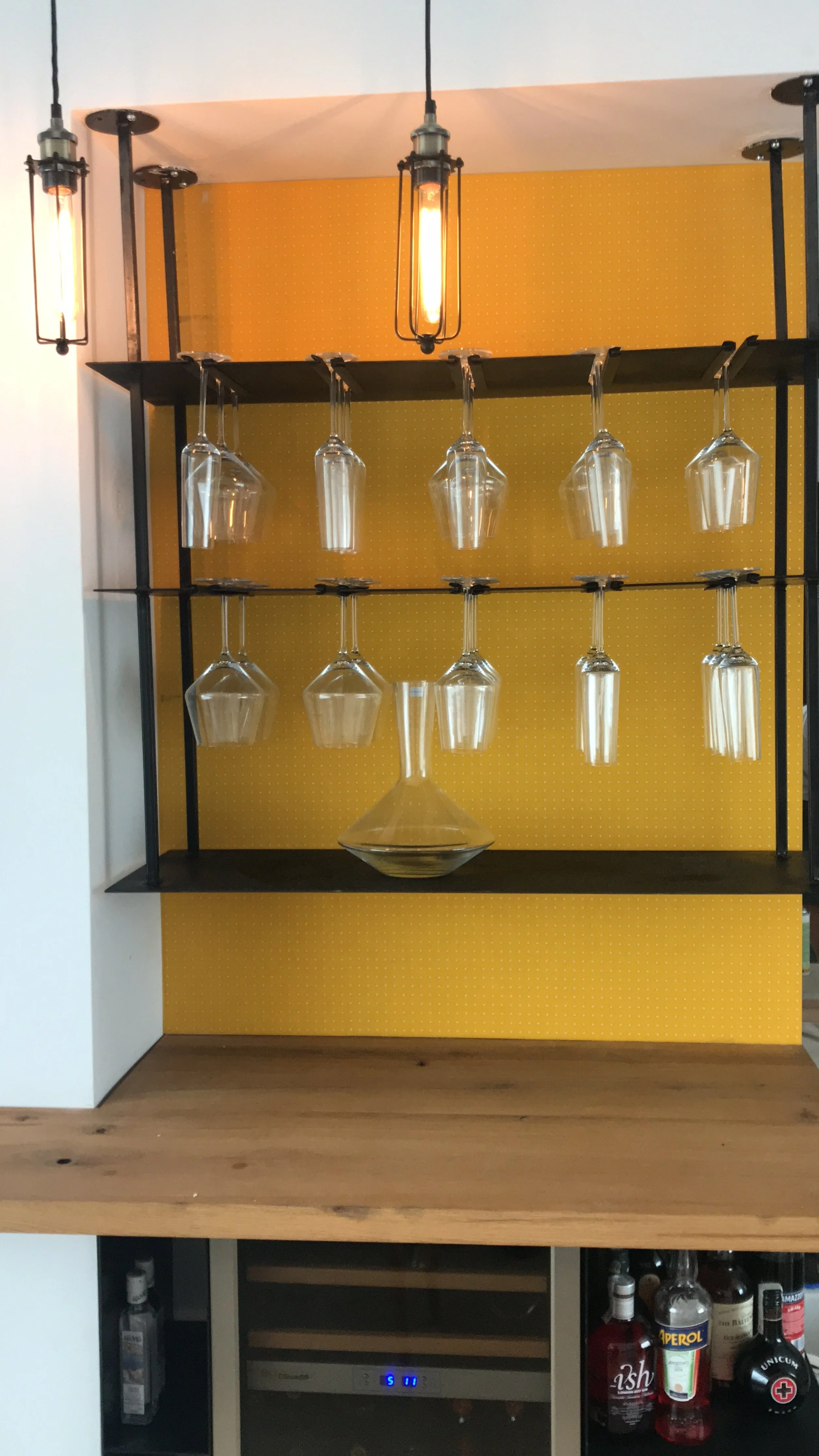 Hanging wine glasses on a rack above a bar counter with a yellow background and industrial light fixtures.