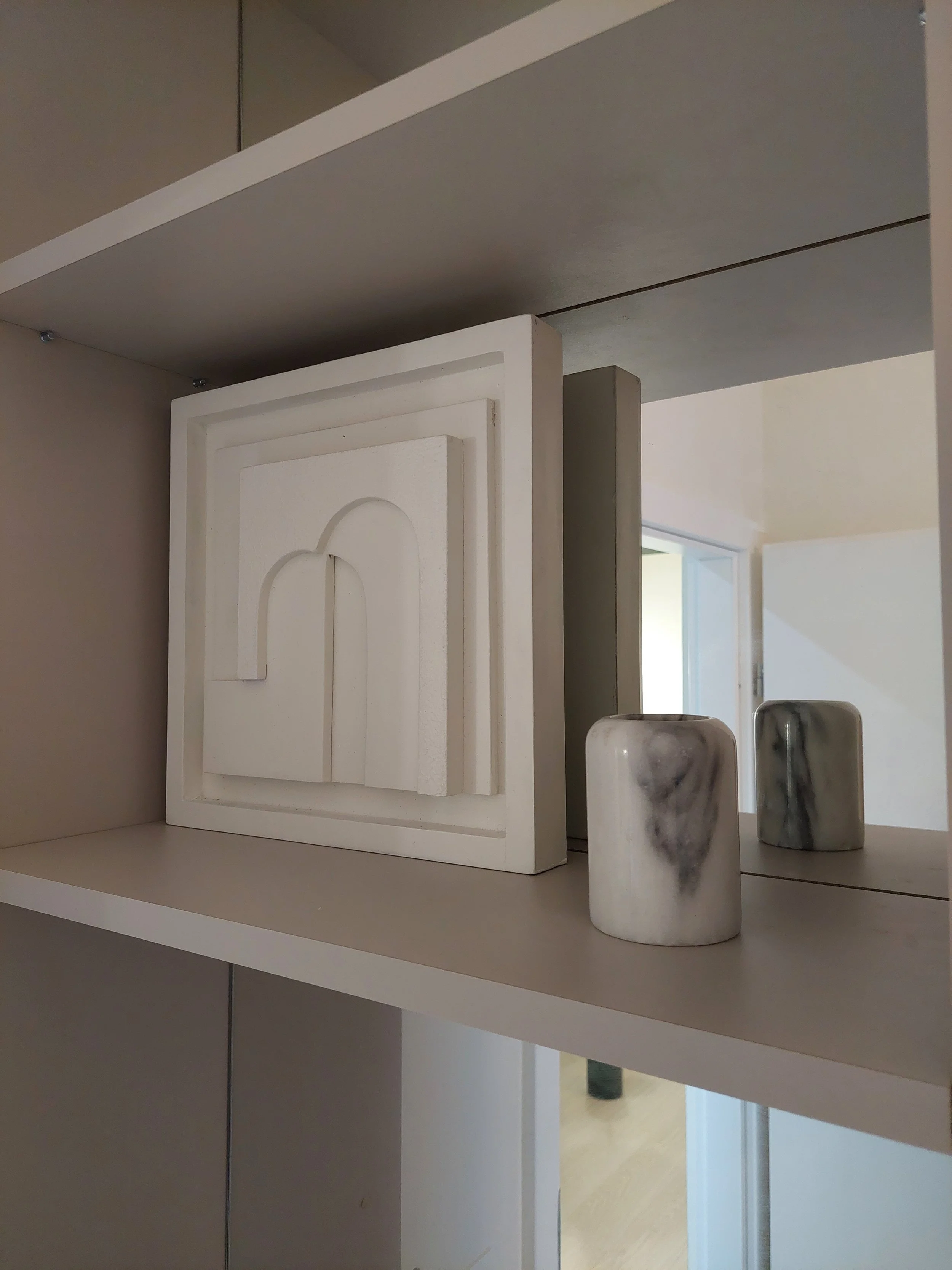 Decorative white 3D wall art and two marble vases on a beige shelf with a mirror behind.