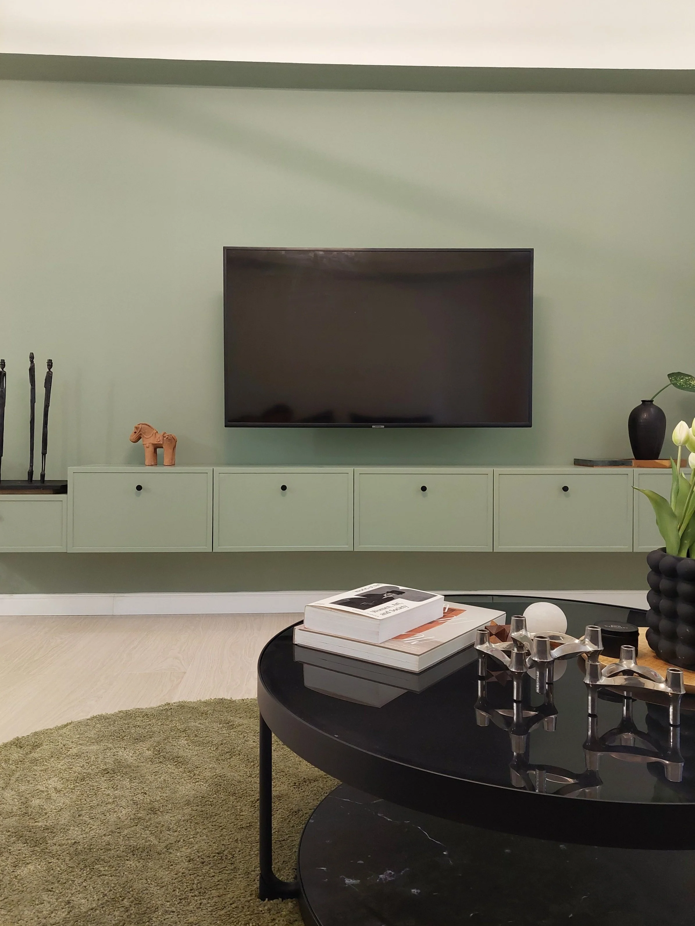 Living room with a mounted TV on a green wall, a black round coffee table with books and decorative objects, and a green rug.