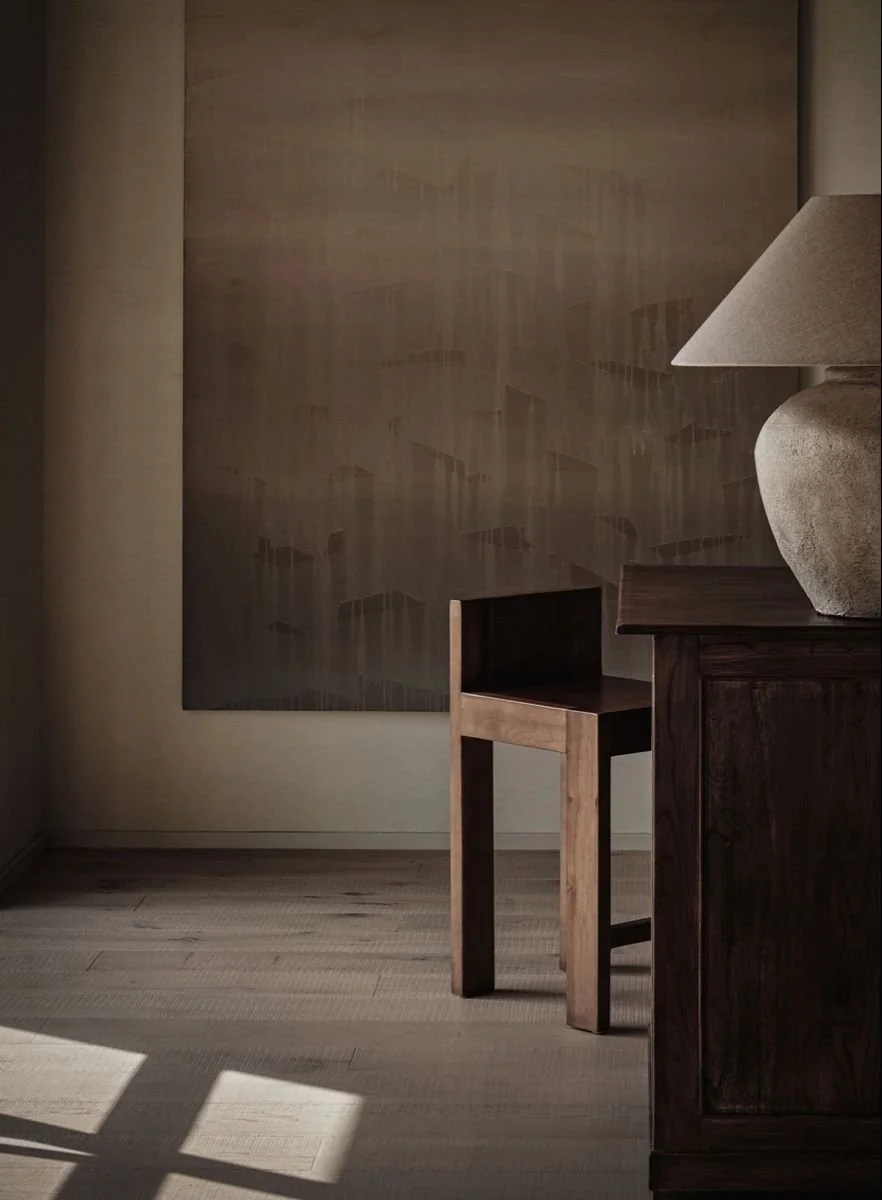 A corner of a room with wooden furniture, including a lamp on a dark wood cabinet, a small wooden table, and a large abstract painting on the wall. Sunlight creates shadows on the light-colored wooden floor.