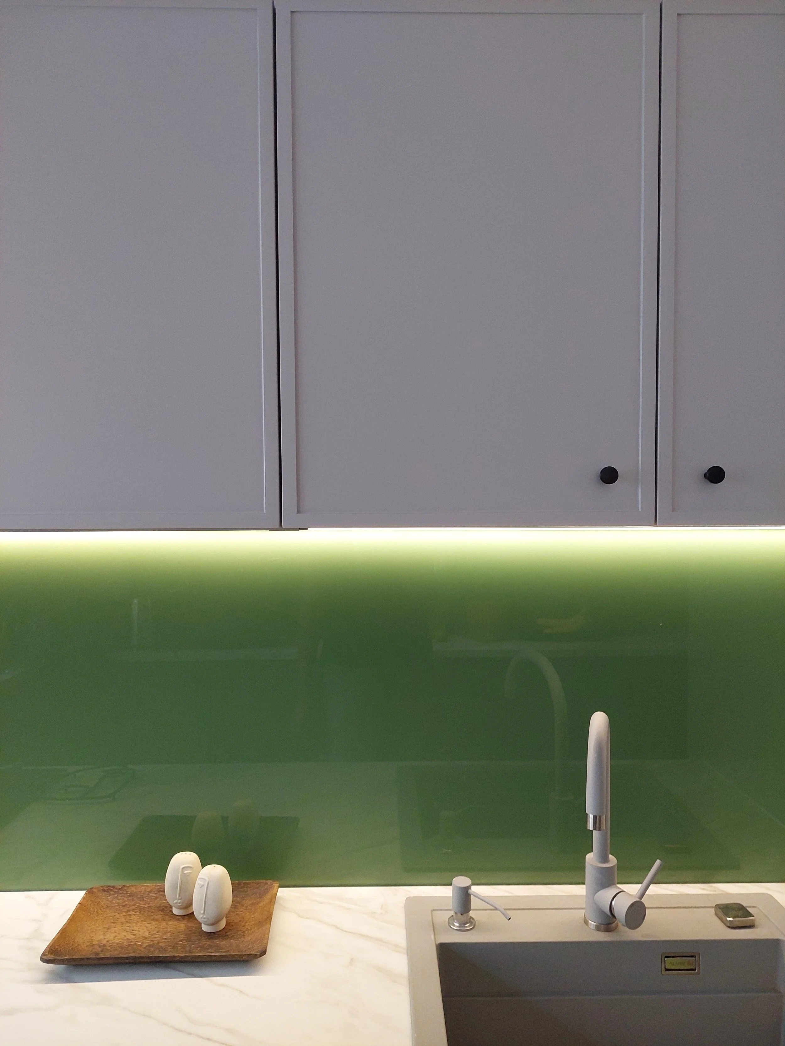 A modern kitchen with white cabinets, a green glass backsplash, a white marble countertop, and a sink with a tall faucet.