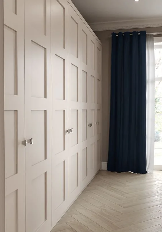 White built-in closet with paneled doors and small silver knobs next to a window with navy blue curtains and sheer white drapes, with light hardwood floor.