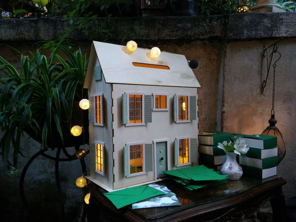 A decorative miniature house with lit windows, surrounded by a string of yellow lights, green envelopes, a small vase with white flowers, and boxes, set on a dark wooden table outdoors at dusk.