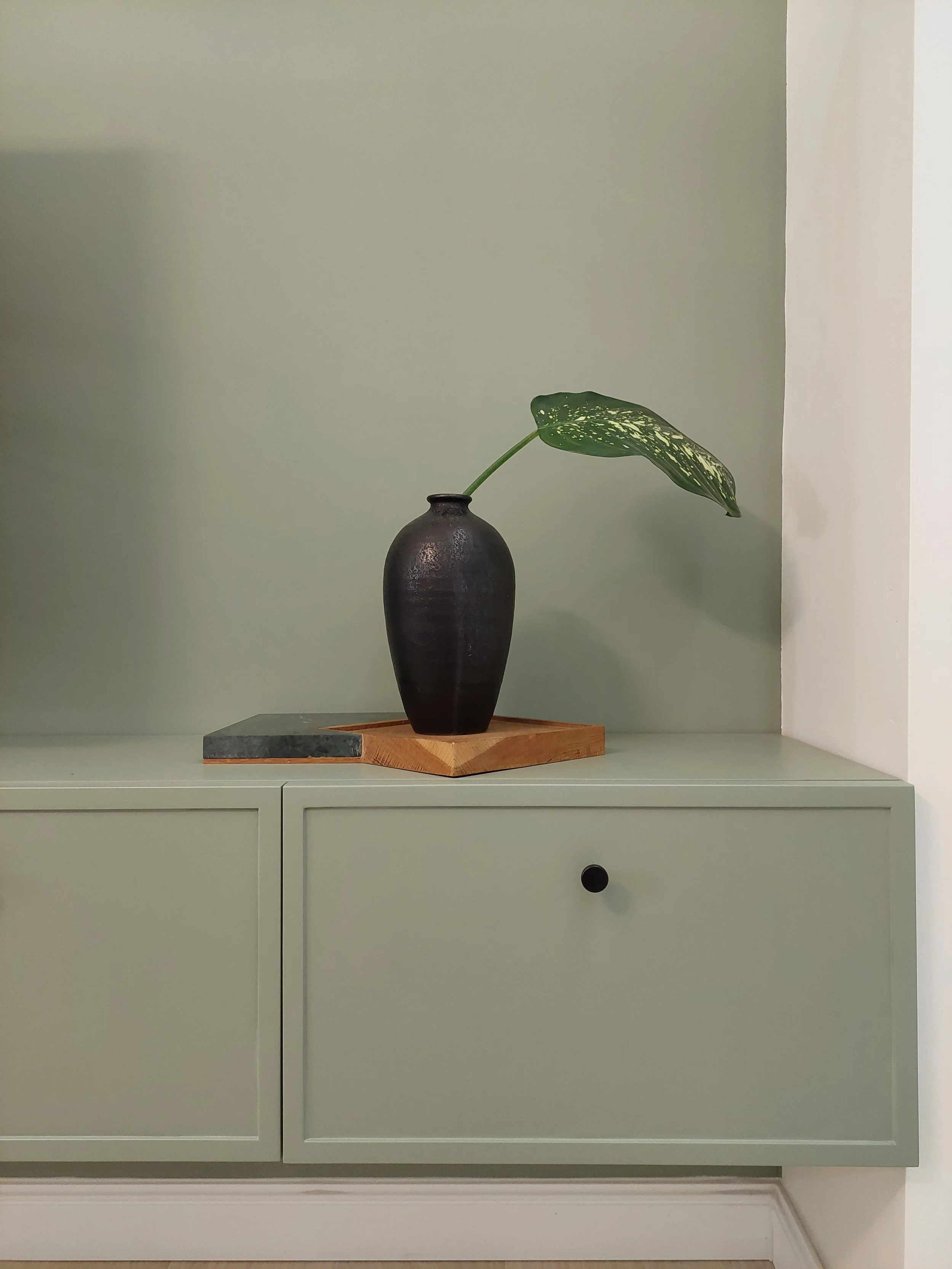 A black vase with a single green leaf, displayed on a wooden and marble platform on top of a light green cabinet against a plain wall.
