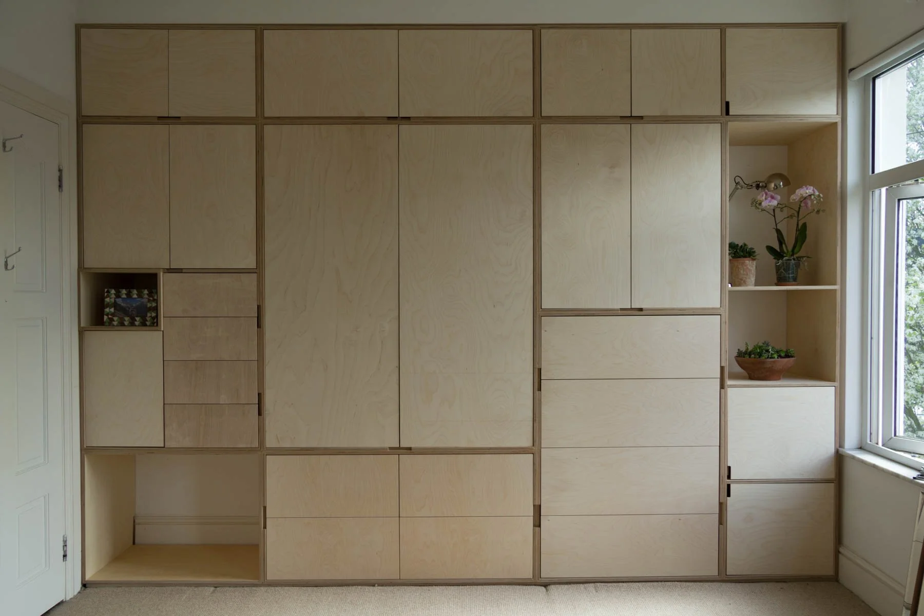 Wall of customized light wood cabinets and shelves in a room with a window showing trees outside.