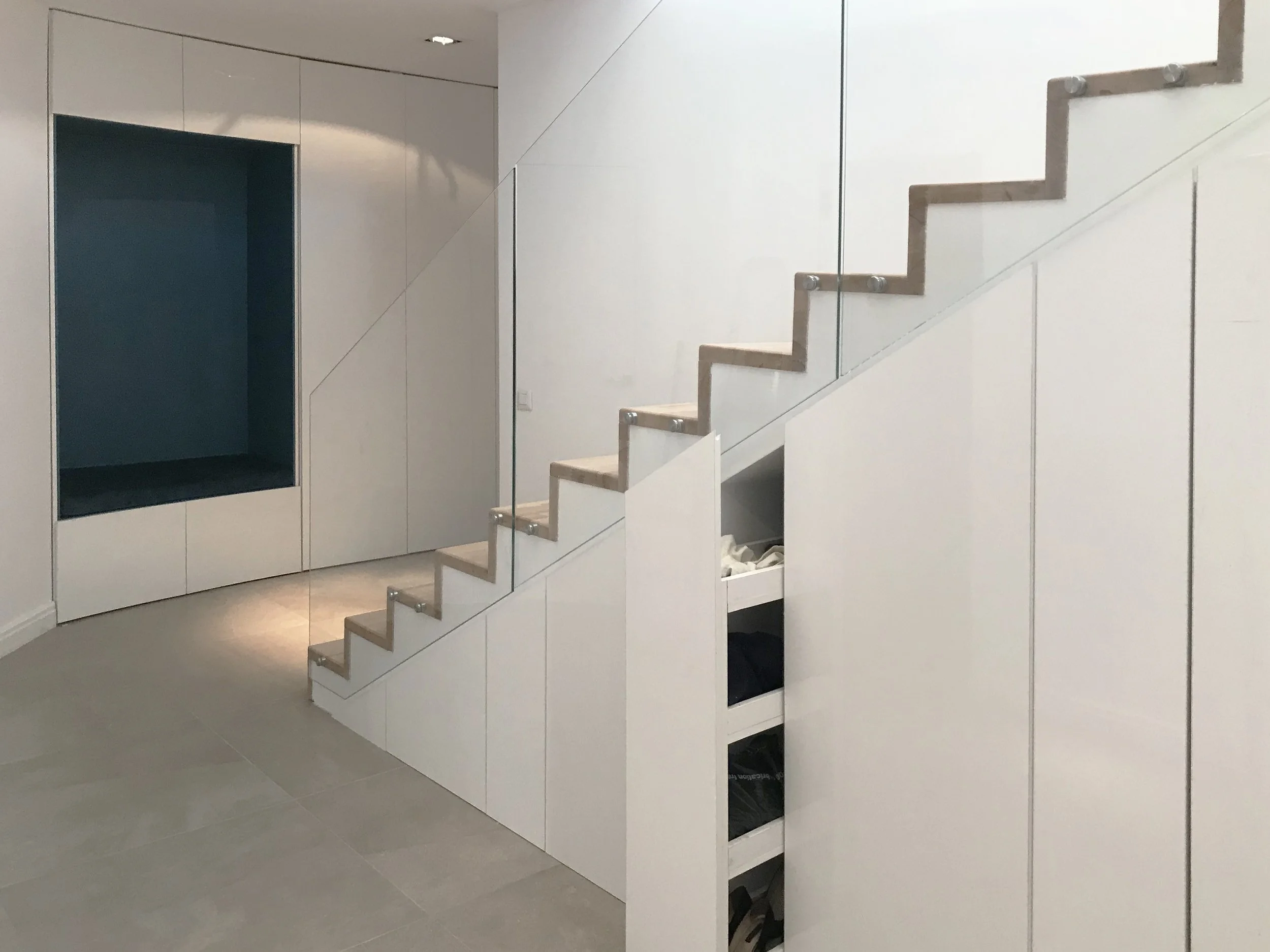 Modern indoor staircase with beige steps, glass railings, and built-in storage underneath, in a minimalist white-walled room.