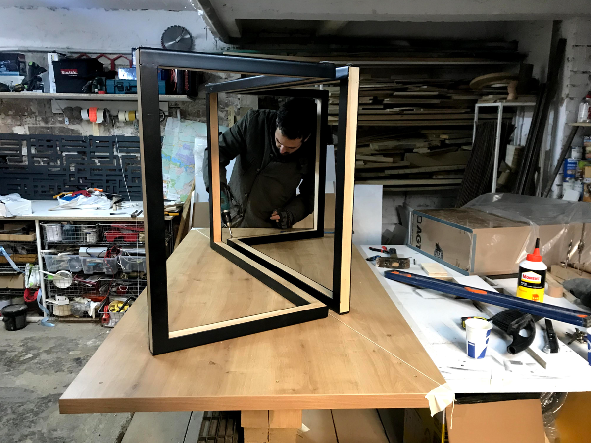 A person working on assembling a black and wood frame structure on a wooden workbench in a workshop.