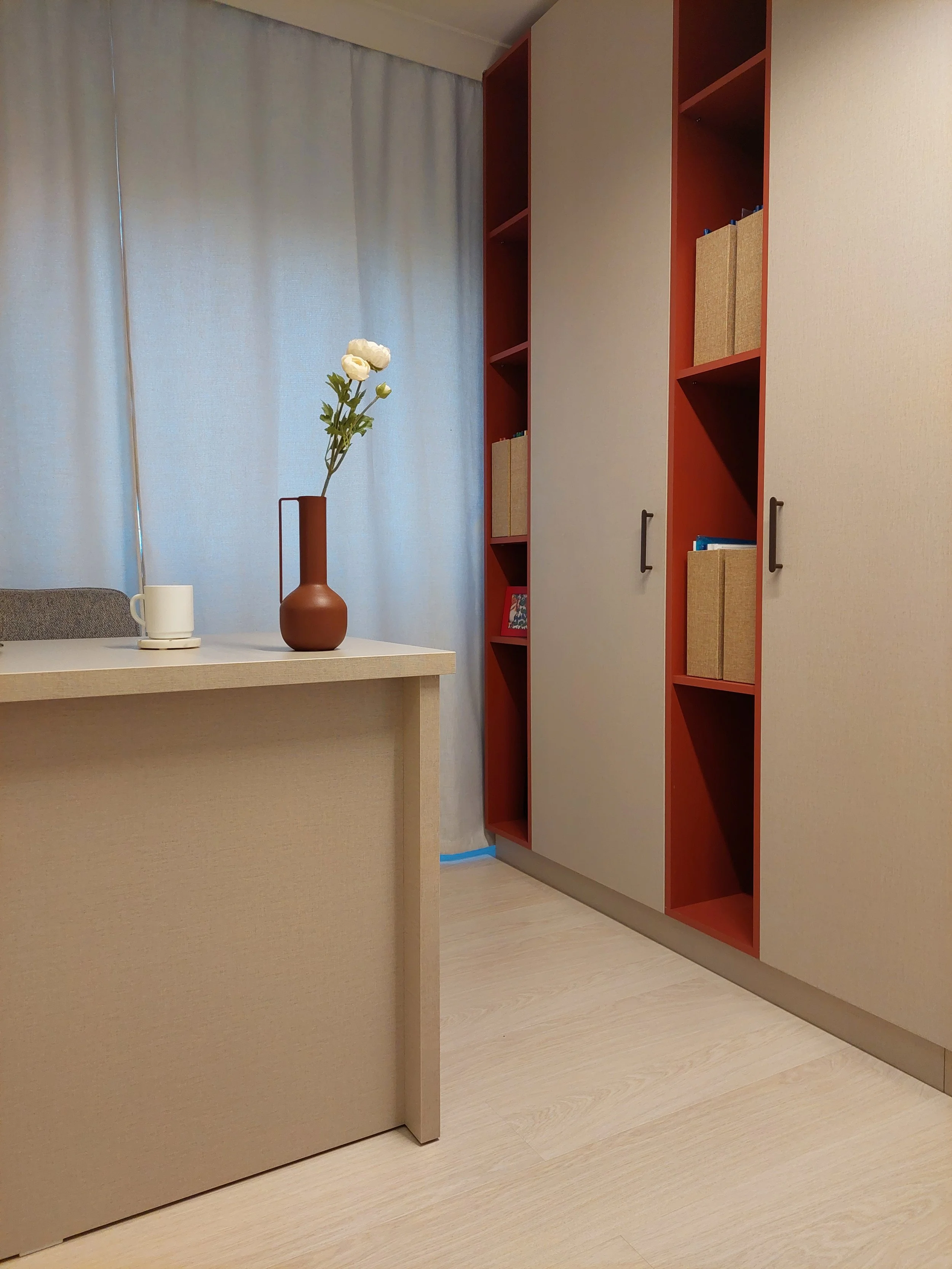 Interior of a room with a beige desk, a brown vase with white flowers, a white mug, a bookshelf with beige and blue books, beige curtains, and a beige wall.