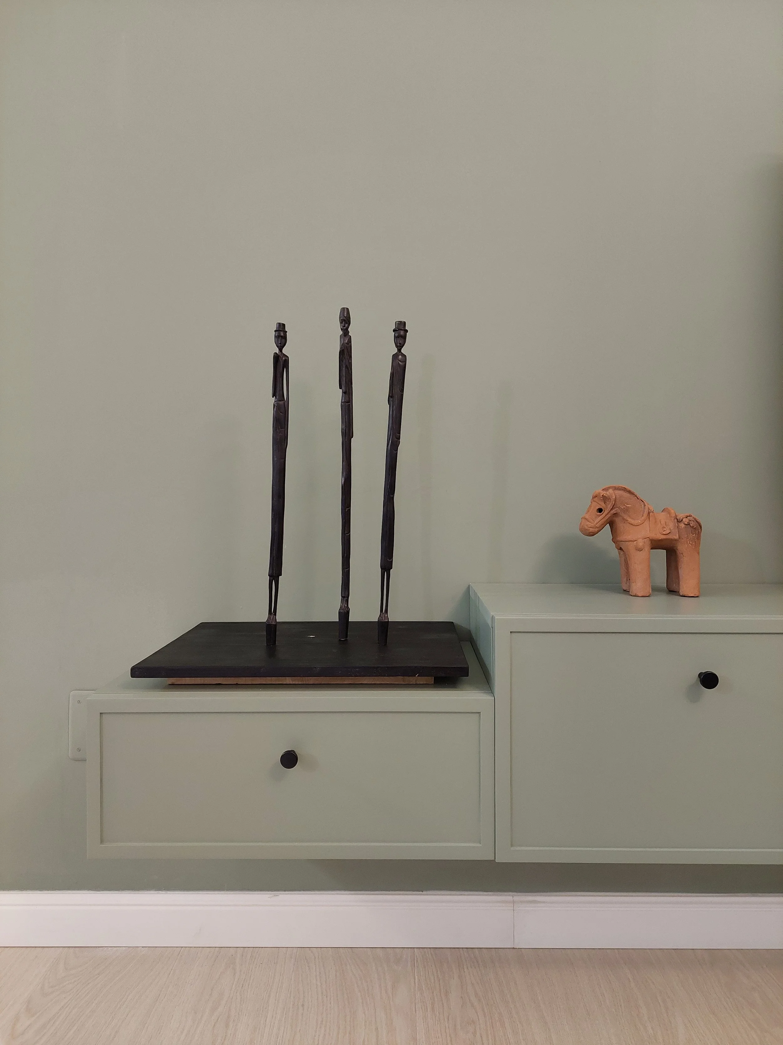 Display of three tall, thin, dark wooden sculptures of elongated human figures on a black platform, and a small terracotta horse figurine on a white cabinet.