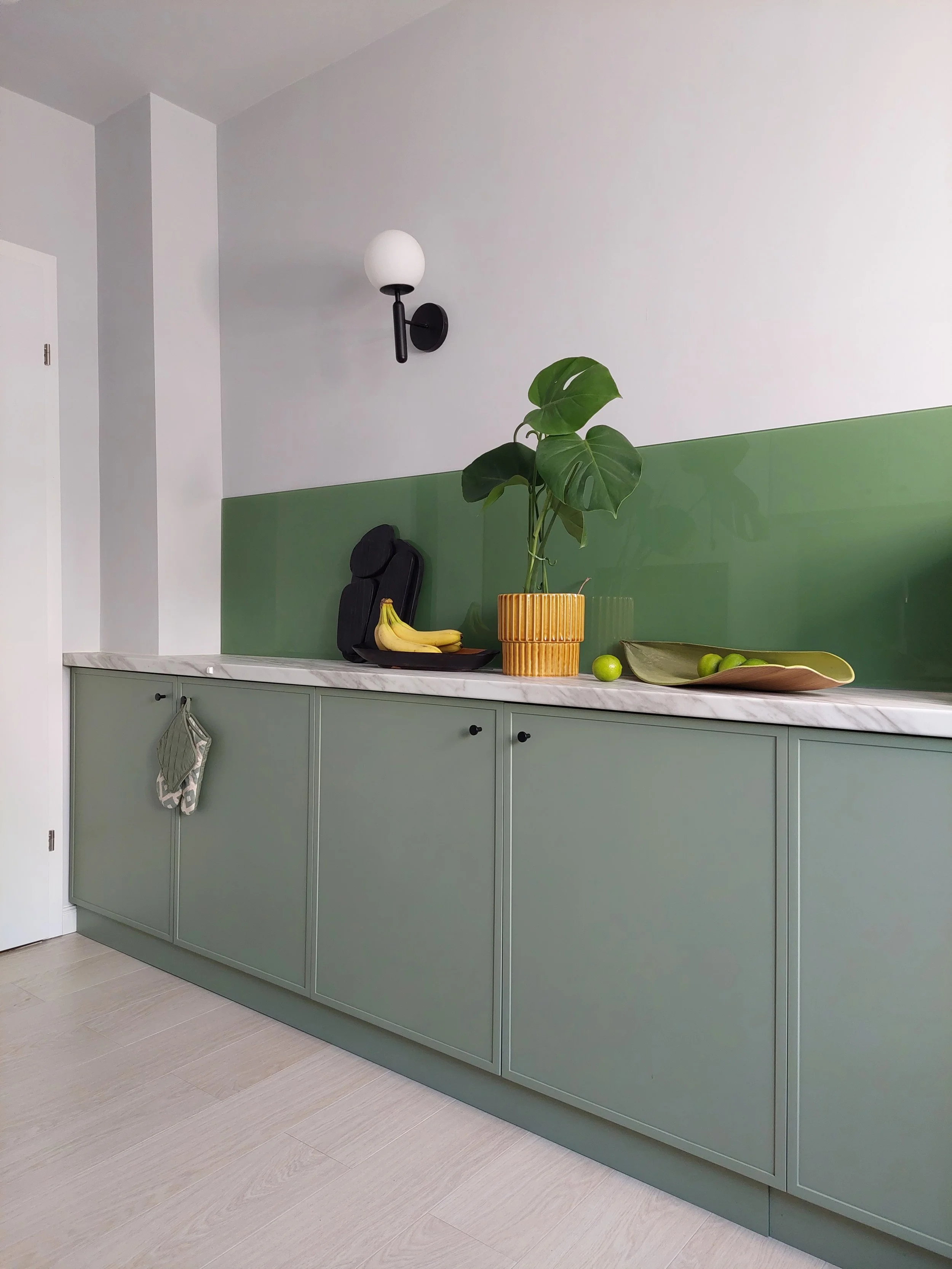 Kitchen counter with green cabinets, marble countertop, wall with green glass backsplash, potted plant, bananas, limes, and wall sconce light.