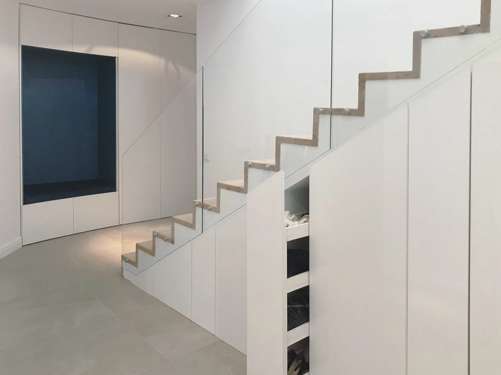 Interior of a modern house with white walls and a staircase with beige steps and glass railing.