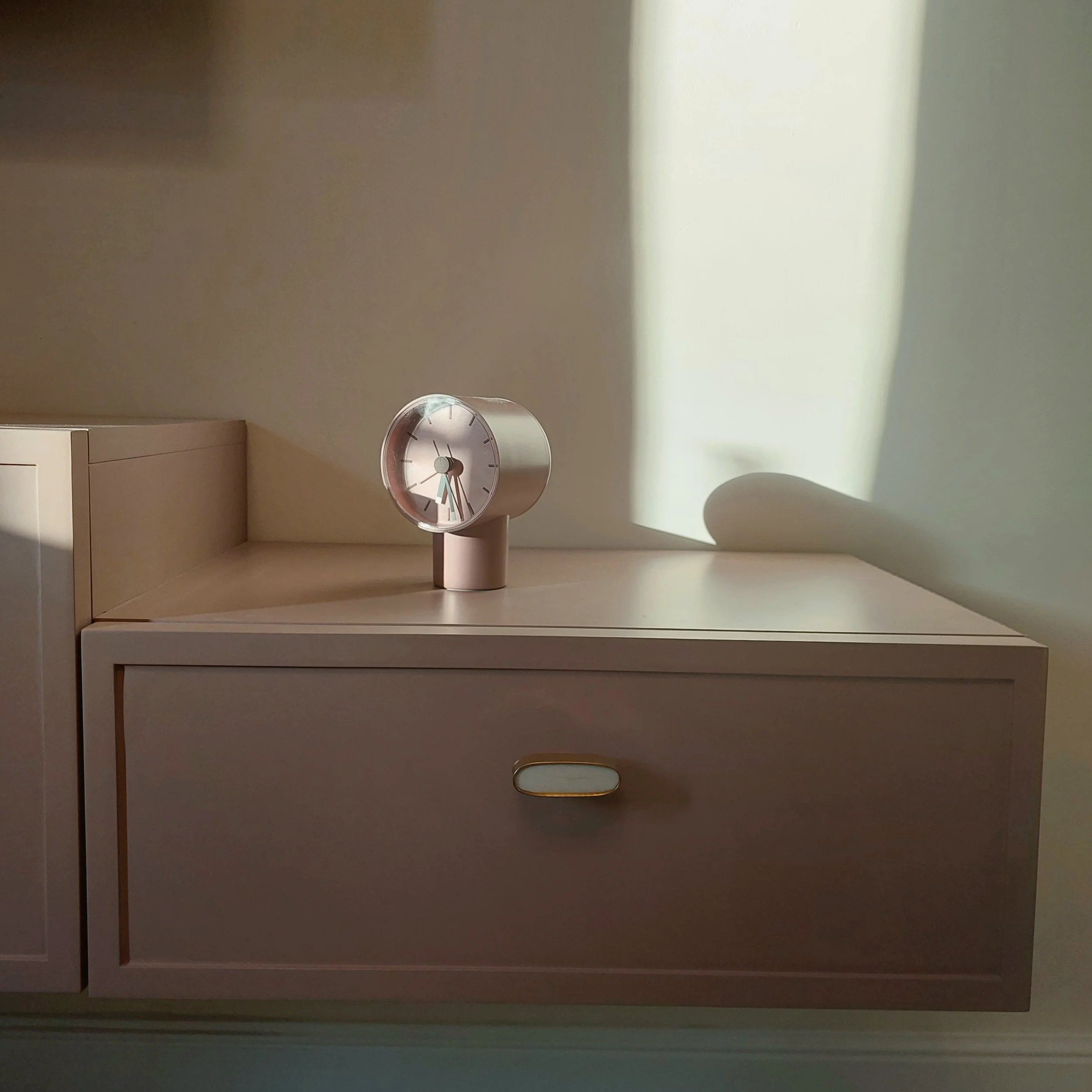 A white and pink round clock on a pink dresser, with shadows on the wall behind.