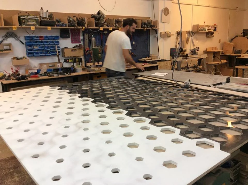 A person working in a workshop with a large sheet of material featuring hexagon cutouts, some cutout sections are filled with black material, and the workspace has tools and shelves on the walls.