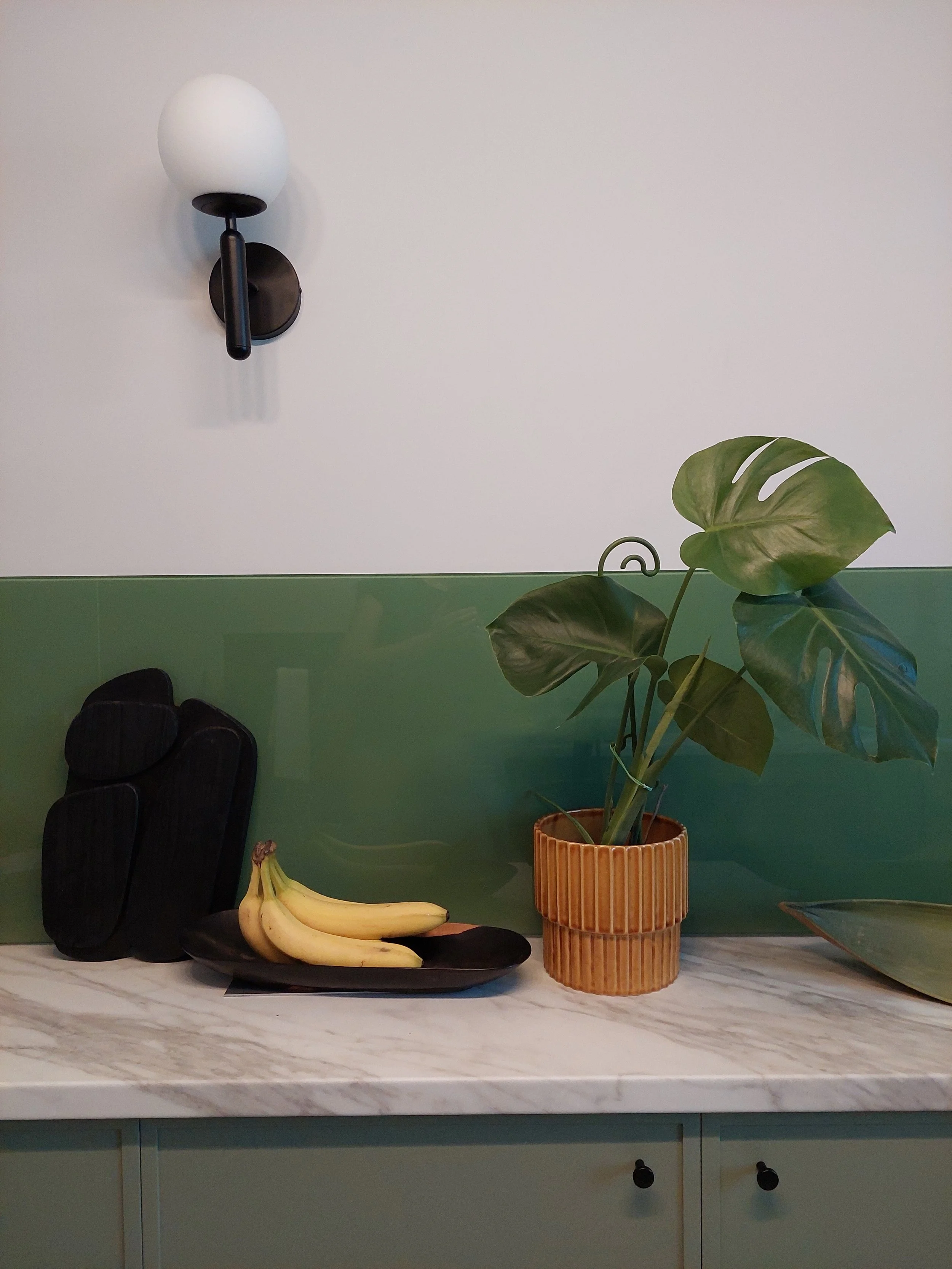 A kitchen countertop with a bunch of bananas on a black plate, a large green leafy plant in a textured brown pot, and black stones arranged behind the plate. A white wall with a green half-wall backsplash and a wall-mounted black and white lamp are visible in the background.