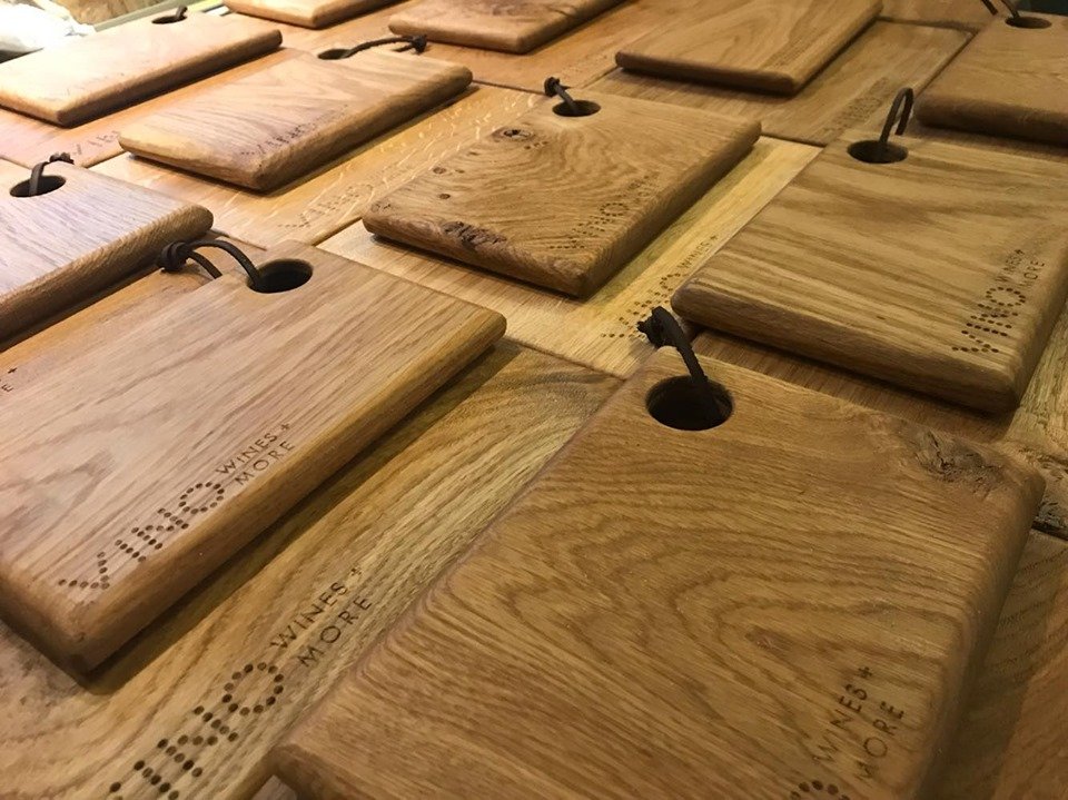 Multiple wooden cutting boards with loops and black cords for hanging, stamped with the 'Wines & More' logo, arranged on a wooden surface.