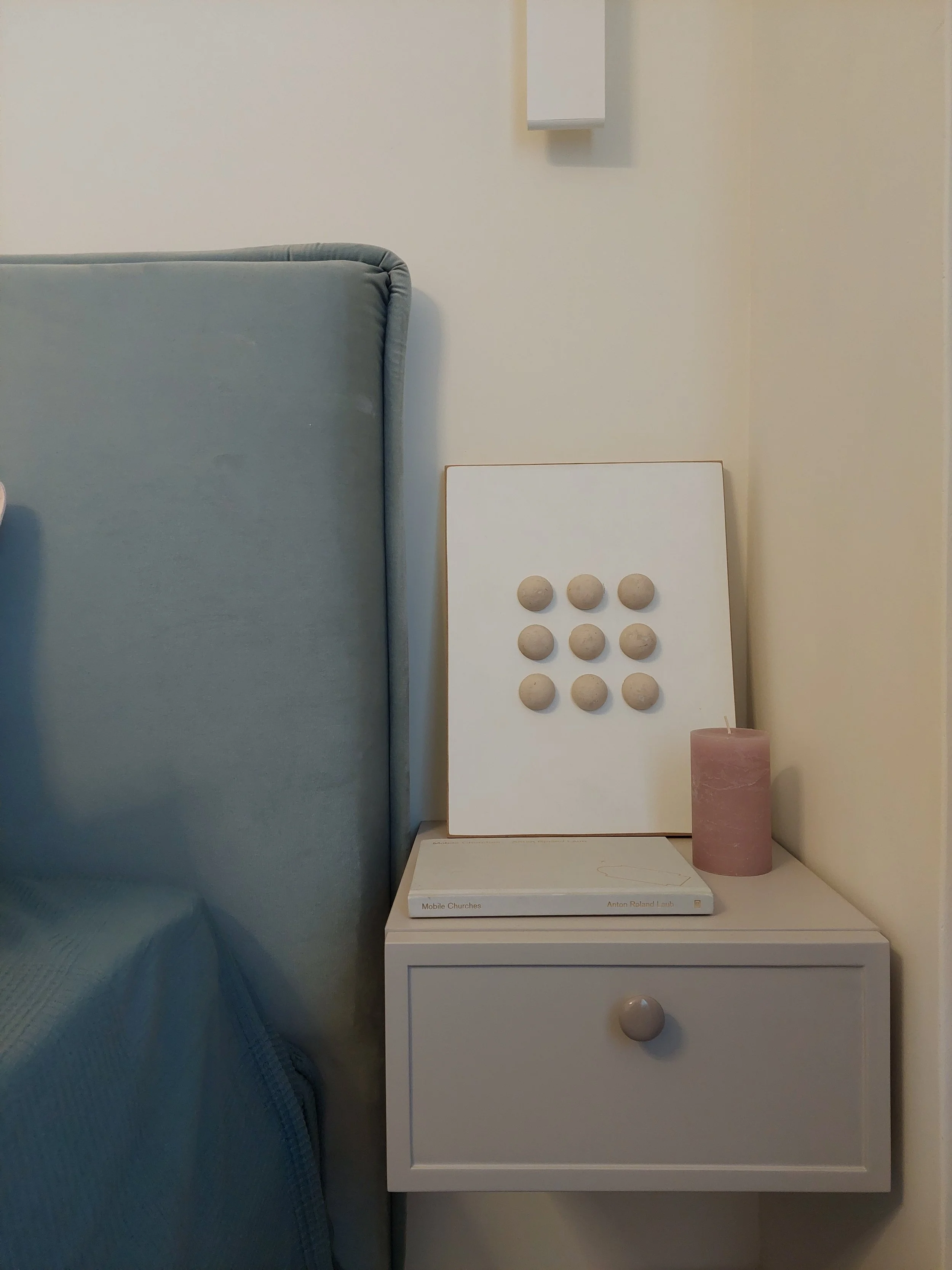 A bedroom nightstand with a candle, a book, and a piece of modern decor art with nine round beige knobs