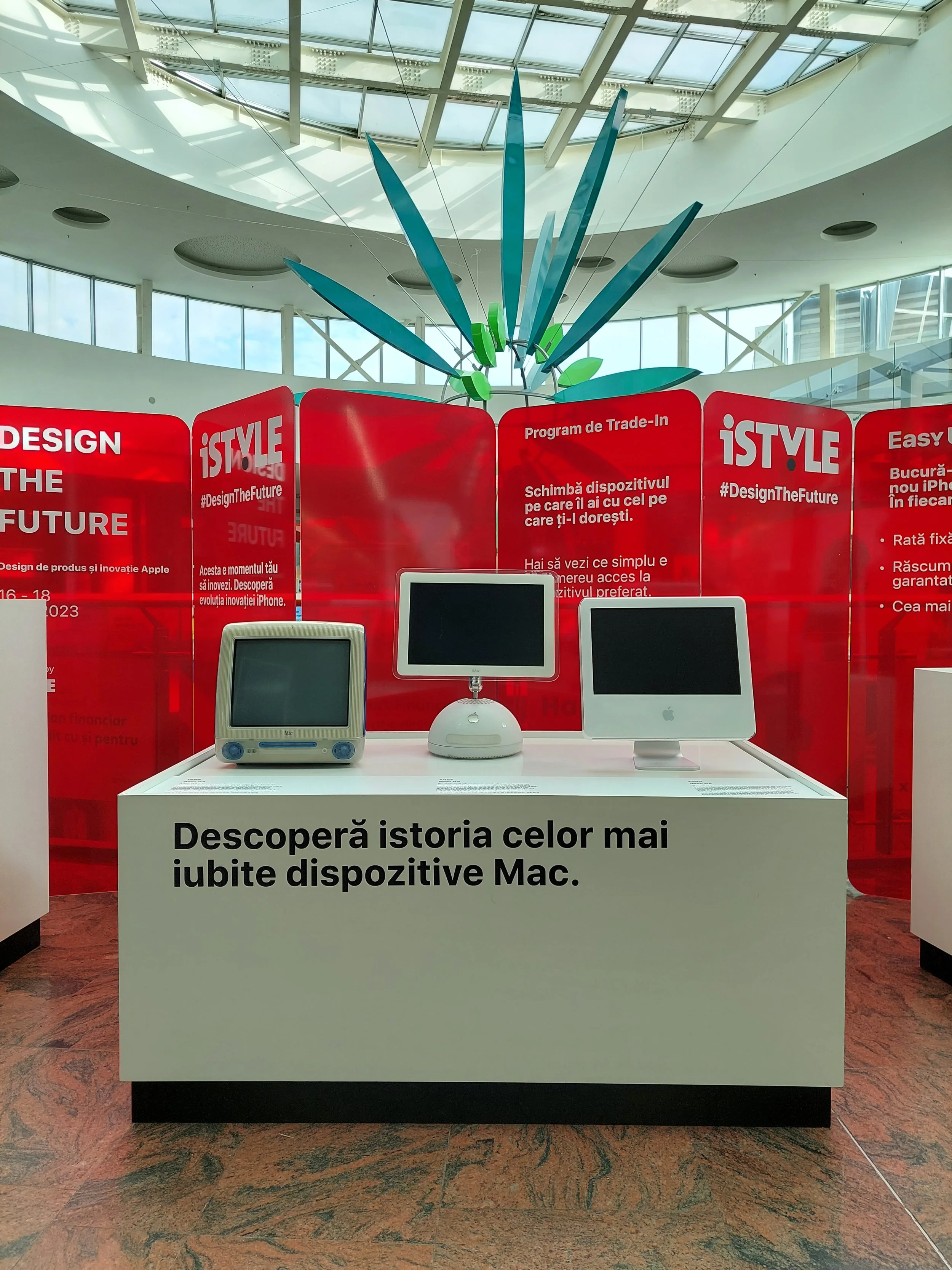 Display of three vintage Mac devices in a museum exhibit, with red informational panels and a modern architectural ceiling overhead.