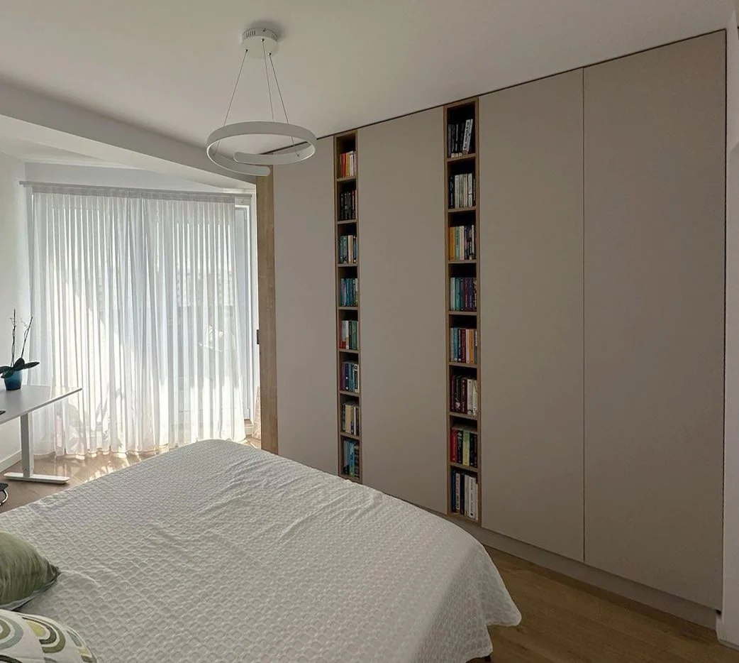A bedroom with a white bedspread, a small window with sheer curtains, a modern ceiling light fixture, a white desk with a potted plant, and a large wardrobe with built-in bookshelves.