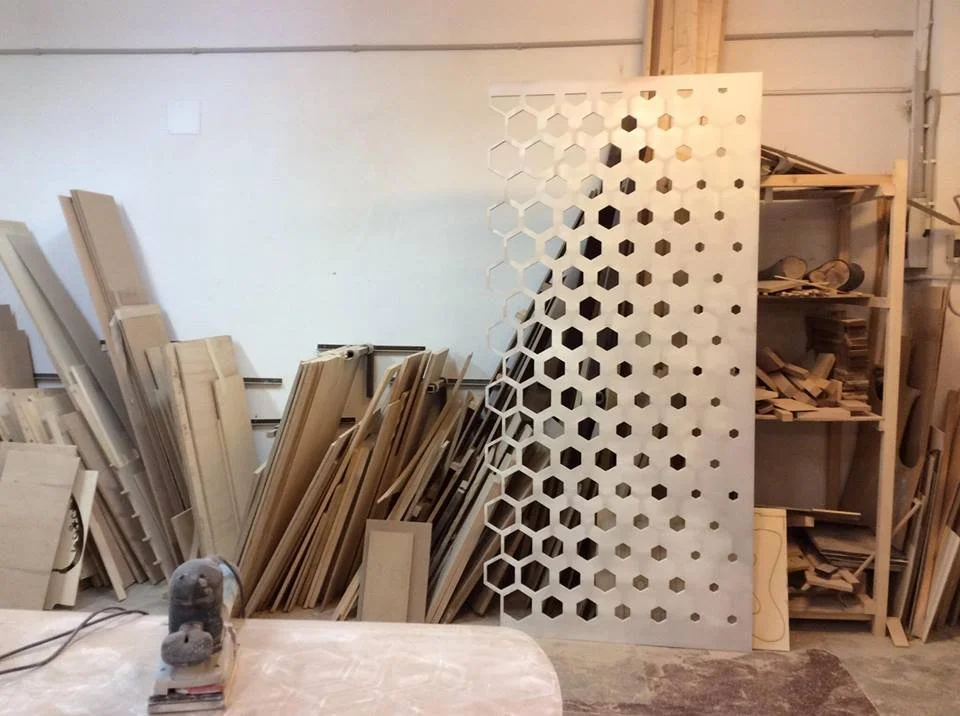 Workshop with woodworking materials including a large wooden panel with hexagon-shaped cutouts, and shelves filled with various pieces of wood.