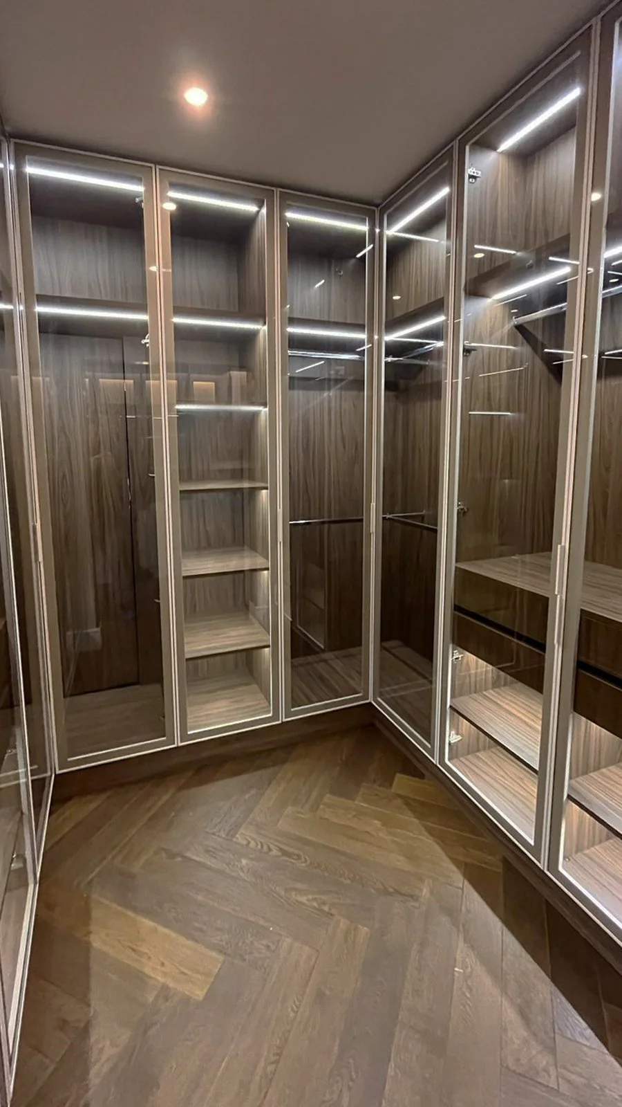 Empty walk-in closet with glass doors, wooden shelves, and drawers, illuminated by ceiling lighting.