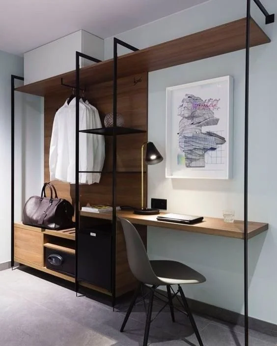 Modern workspace featuring a built-in desk, a gray chair, and a decorative shelving unit with a hanging white shirt and a black bag, against a light-colored wall with framed artwork.