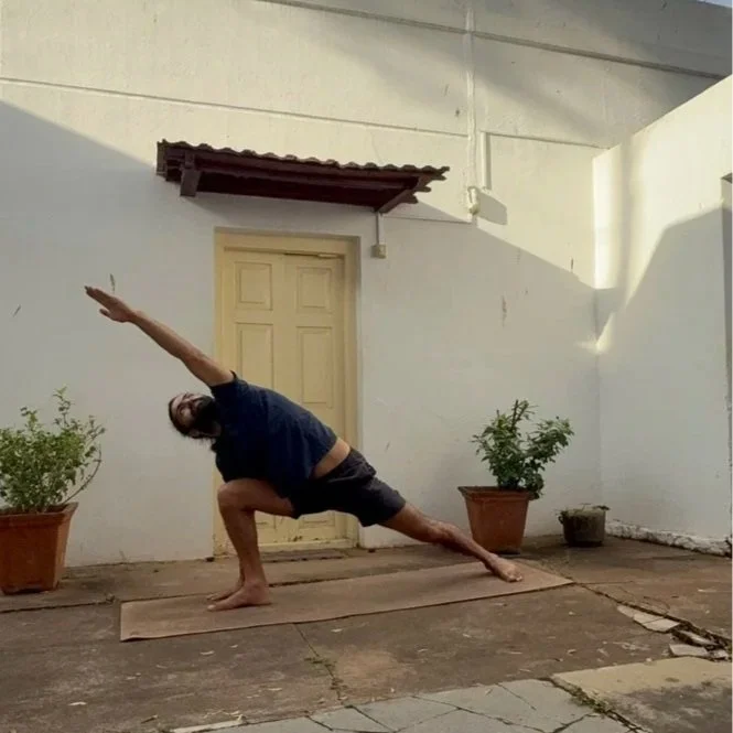 Person practicing yoga outside on a mat, performing a side stretch with one arm extended overhead and the other hand on the ground, in front of a white wall with potted plants.