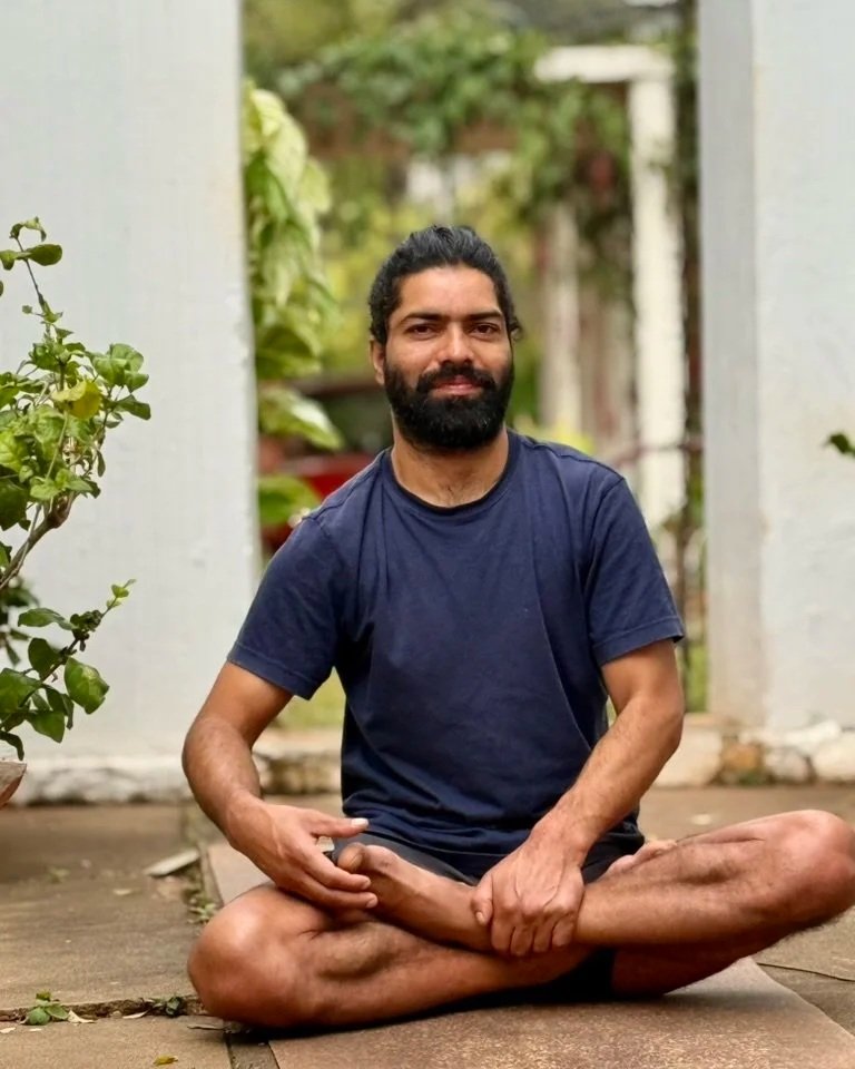 A man with dark hair and a beard sitting cross-legged on a concrete surface outdoors, surrounded by greenery, wearing a navy blue t-shirt.