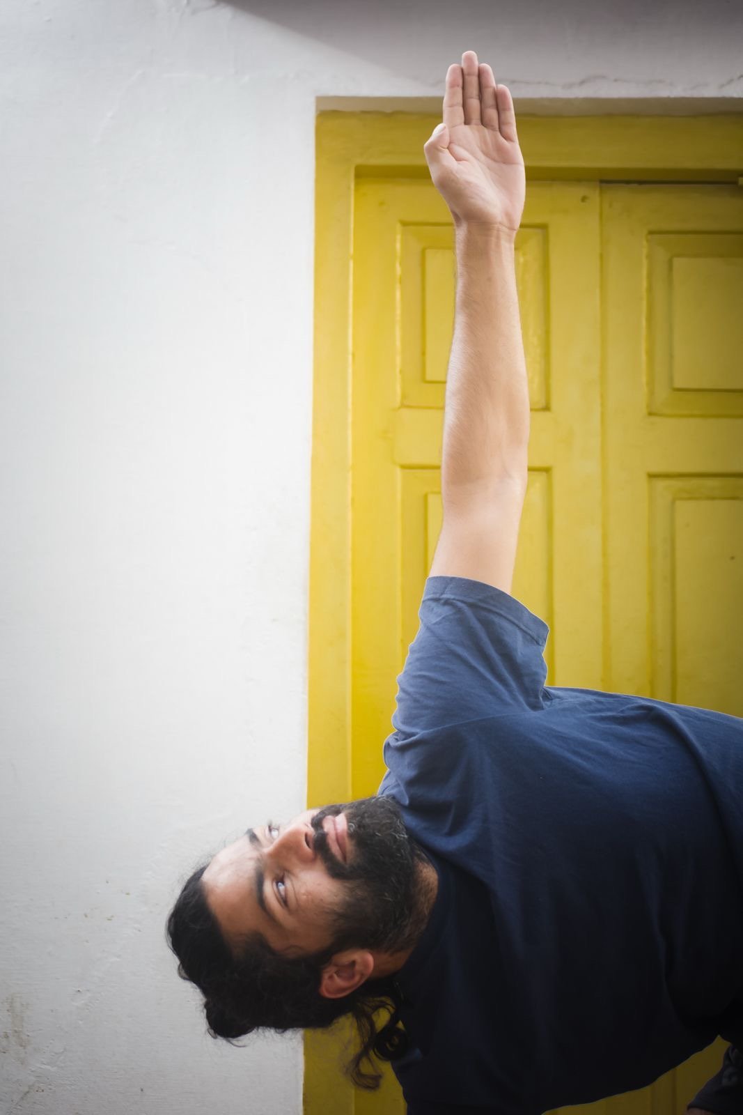 A man lying on the floor with his head turned to the left, stretching his right arm vertically upward, in front of a yellow door with a white wall.