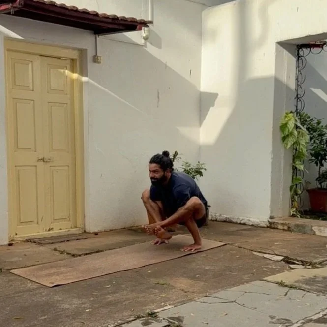 A man practicing yoga outdoors on a mat in front of a white wall and door, with sunlight casting shadows.