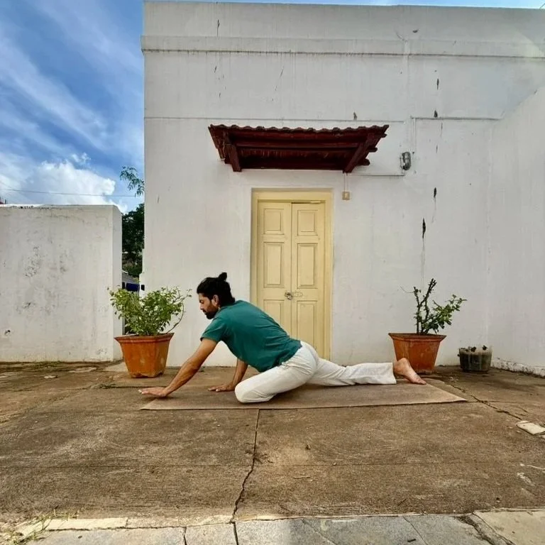 A man practicing yoga outdoors on a mat in front of a white building, surrounded by potted plants.