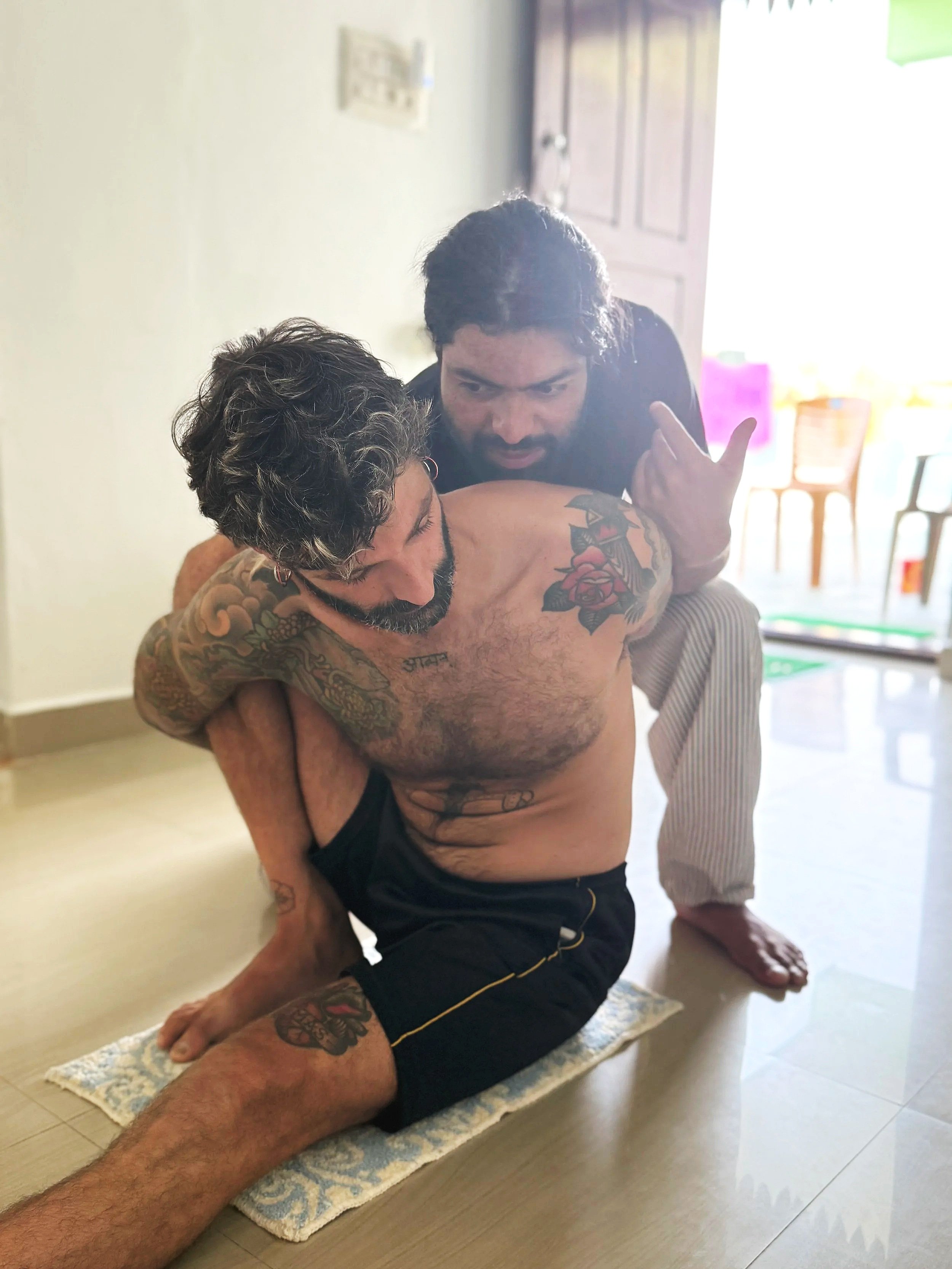 Two men are practicing Brazilian Jiu-Jitsu on a small mat in a home setting. One man is on top, wearing black shorts, with tattoos on his arms and chest, and is gripping his opponent's leg. The other man is on his back, leaning back, wearing a black shirt with white and black striped pants, and is looking intensely at his opponent. The room has a wooden door and chairs in the background.