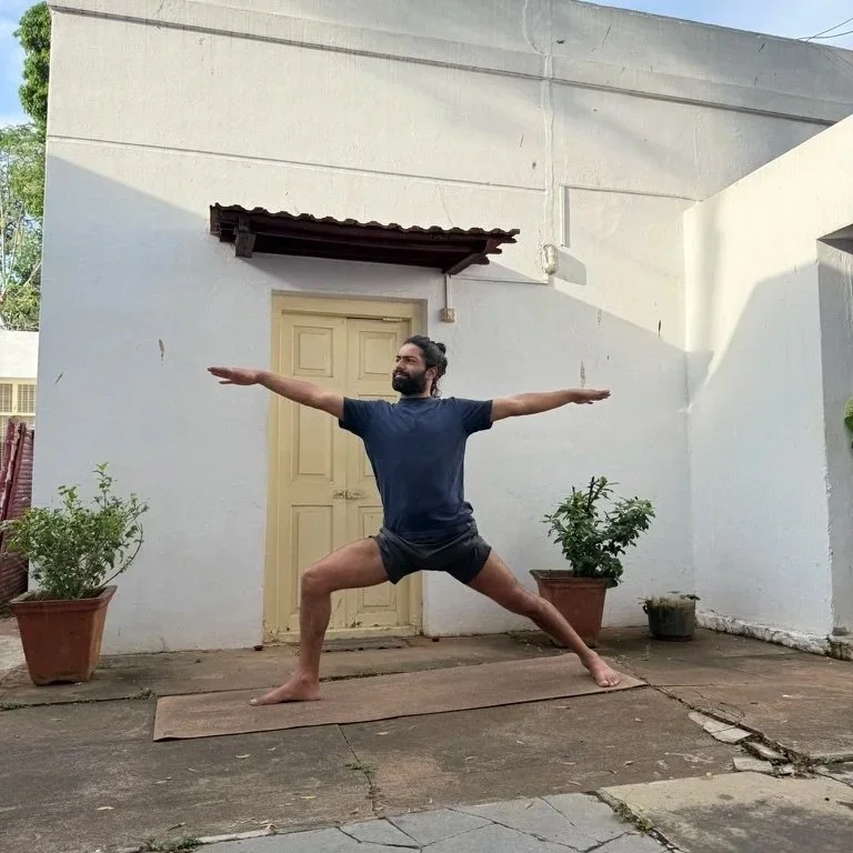 Man practicing yoga outdoors on a mat, performing Warrior II pose with arms extended and legs wide apart in a patio area with potted plants and a white wall.