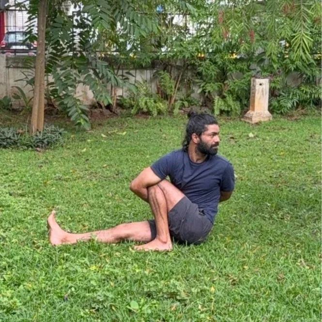 Man outdoors on grass, stretching or yoga pose, with one leg bent and holding it with his arms, in front of green plants and trees.