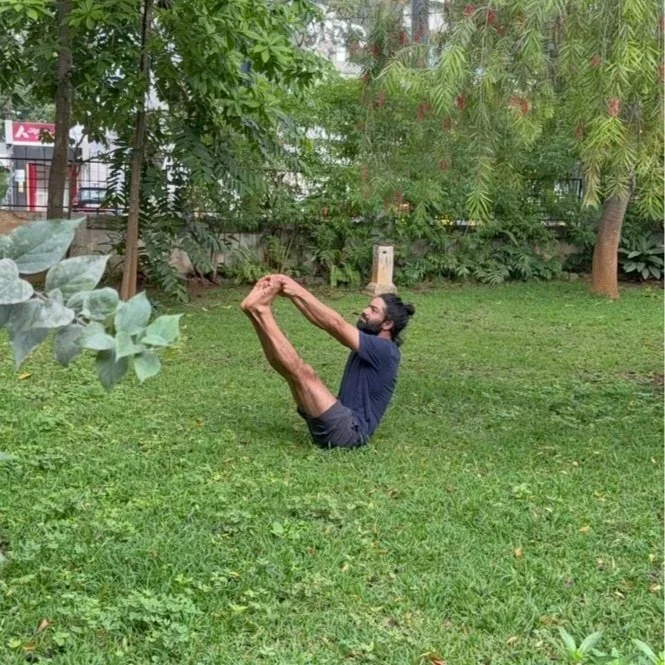A man doing a yoga pose on a grassy area in a park, with trees and bushes in the background.